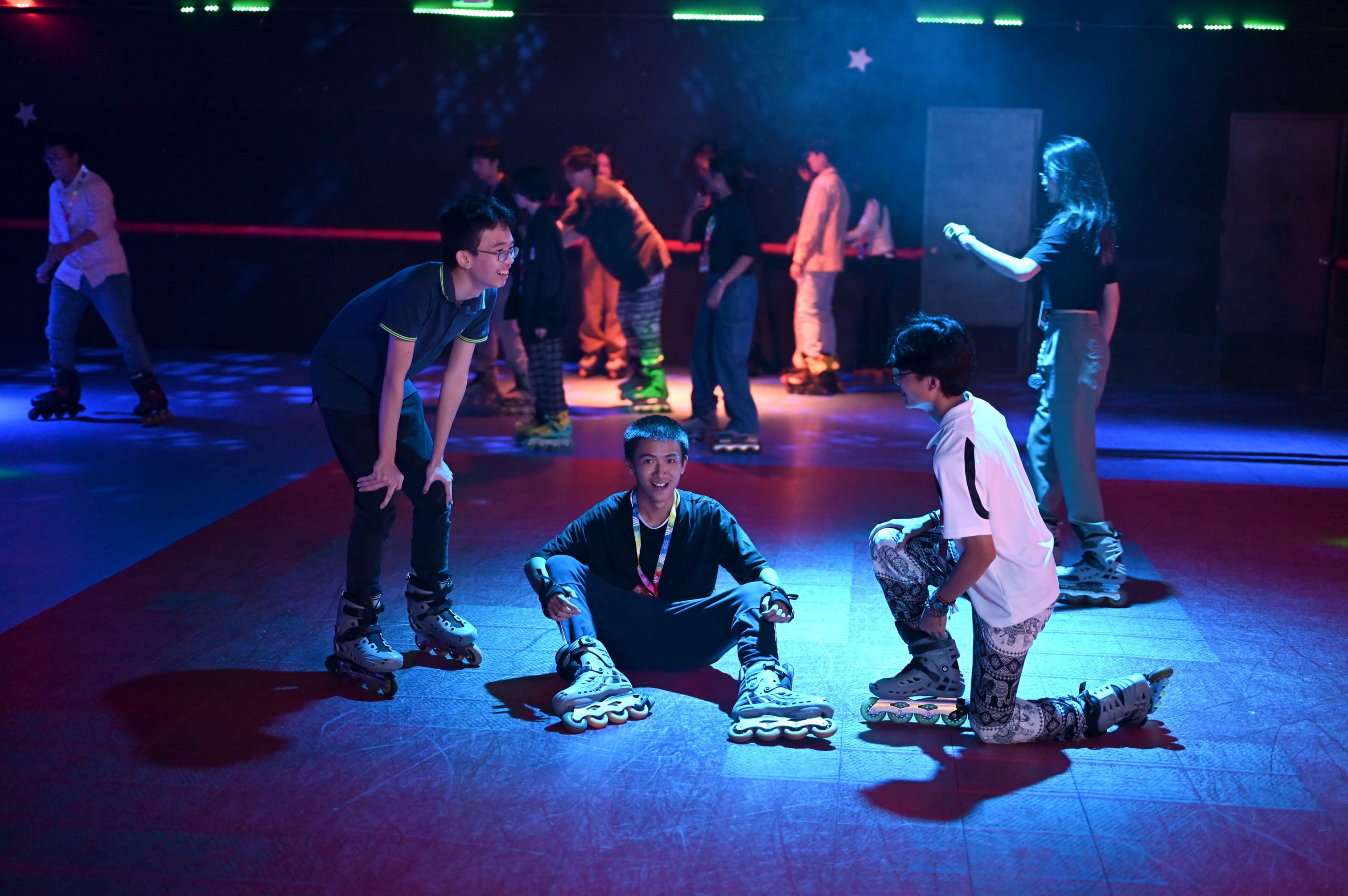 Teens wearing inline skates on a dimly lit roller rink with colorful lights, one sitting on the floor smiling.