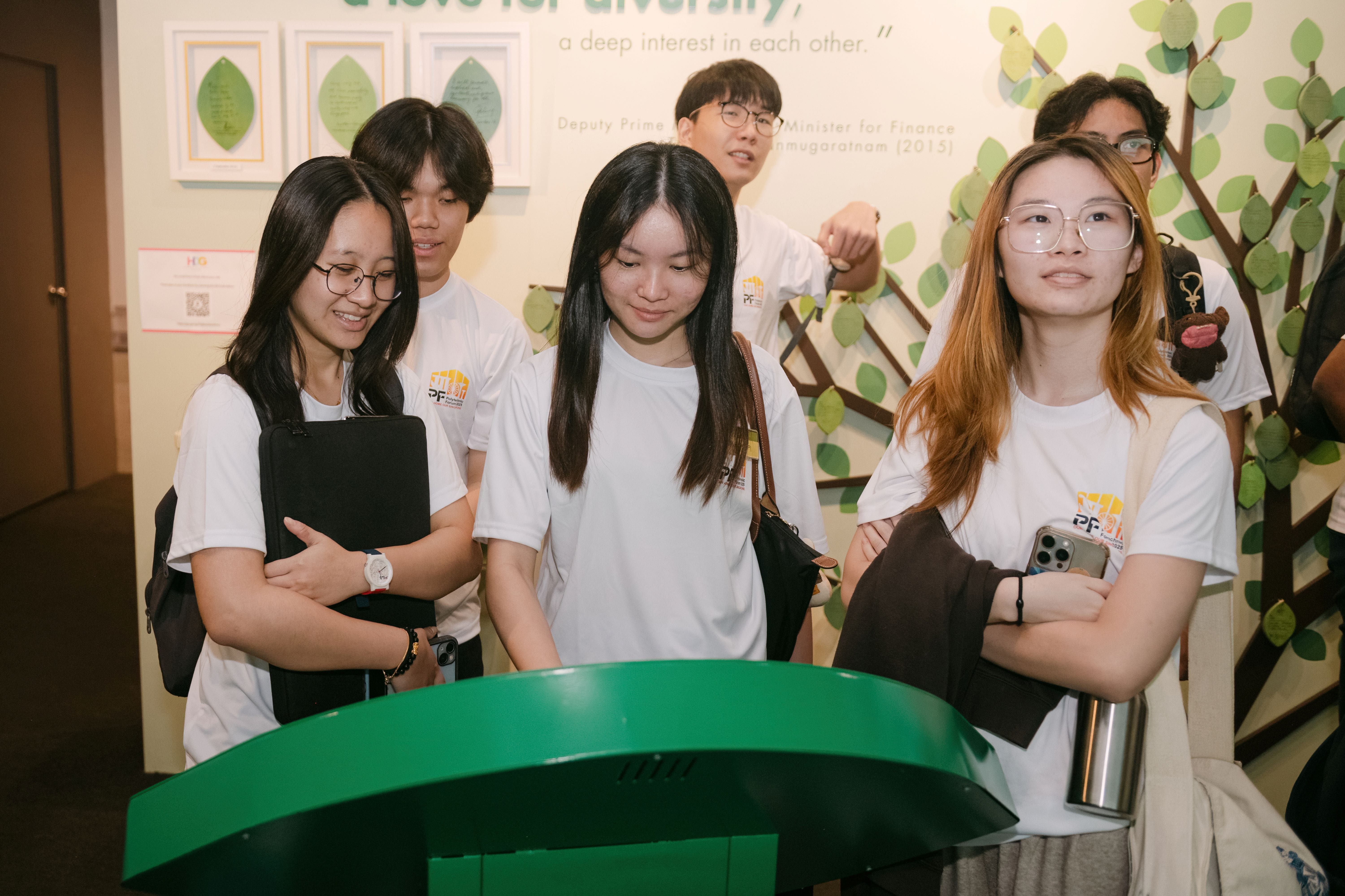 Five young people in matching white T-shirts stand together at a museum exhibit with a decorative tree wall display.