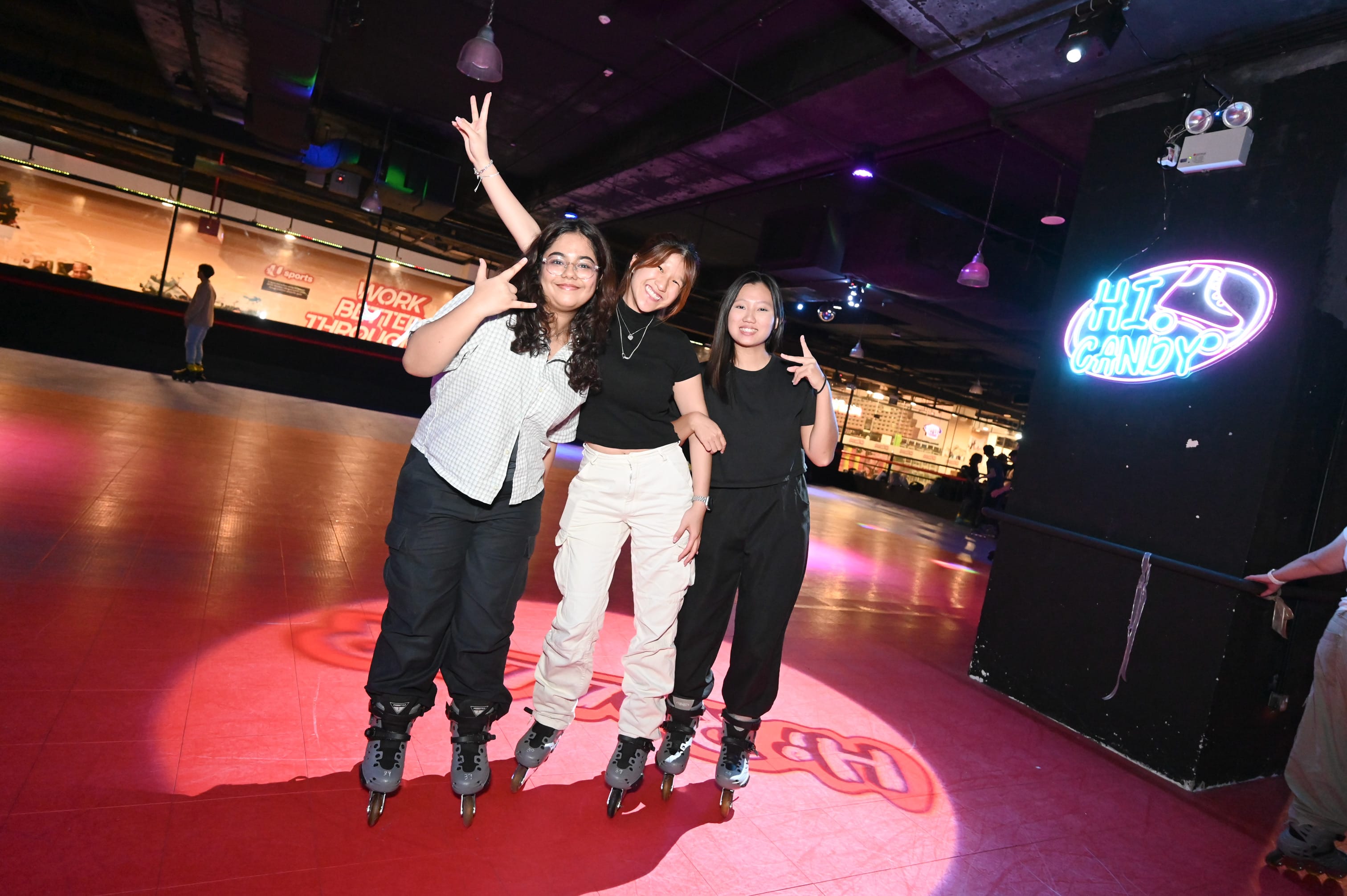 Three young women wearing inline skates pose and smile on a dimly lit roller rink with neon signs.