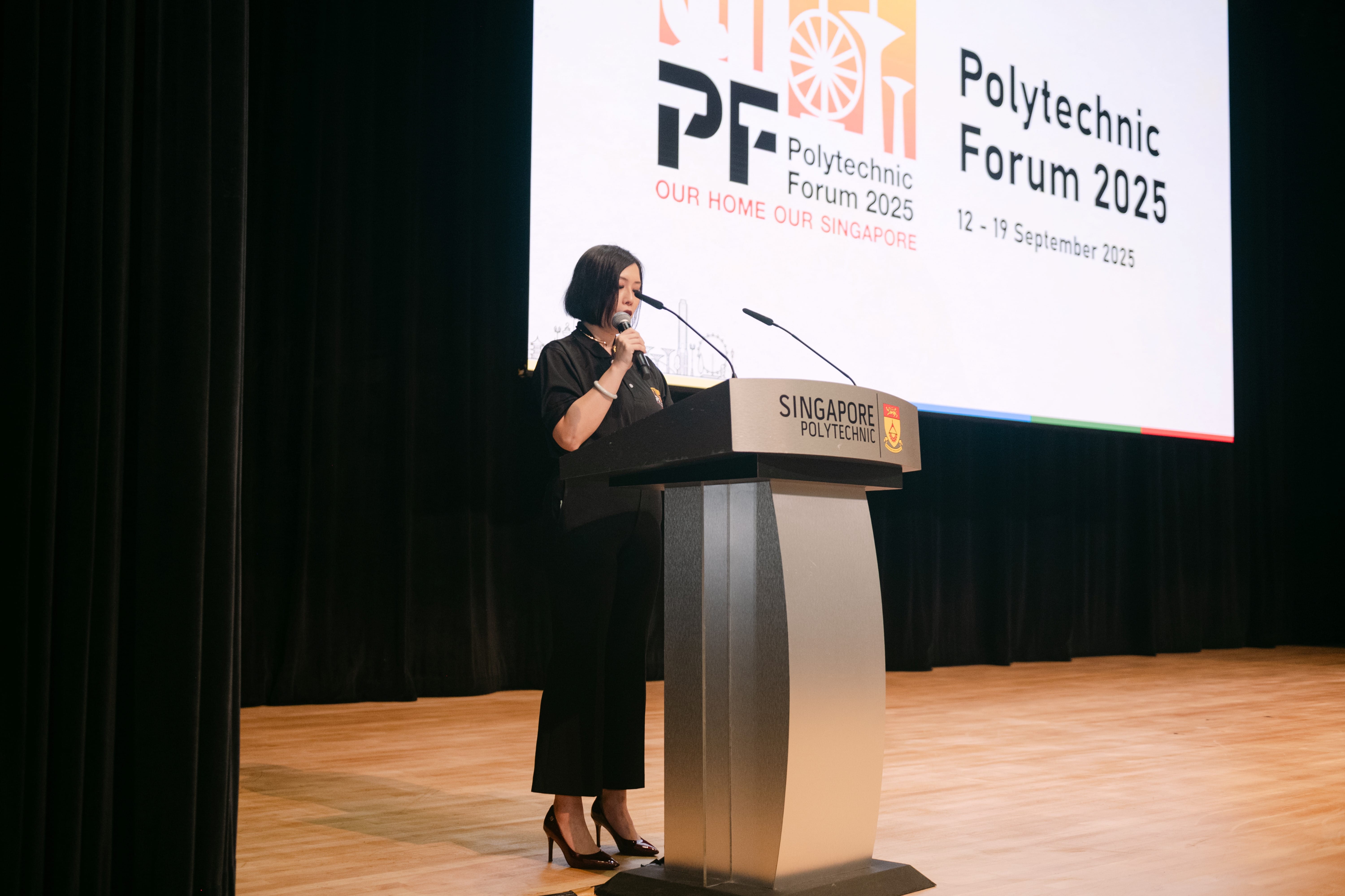 Woman in black speaking at a Singapore Polytechnic podium with a Polytechnic Forum 2025 banner behind her.