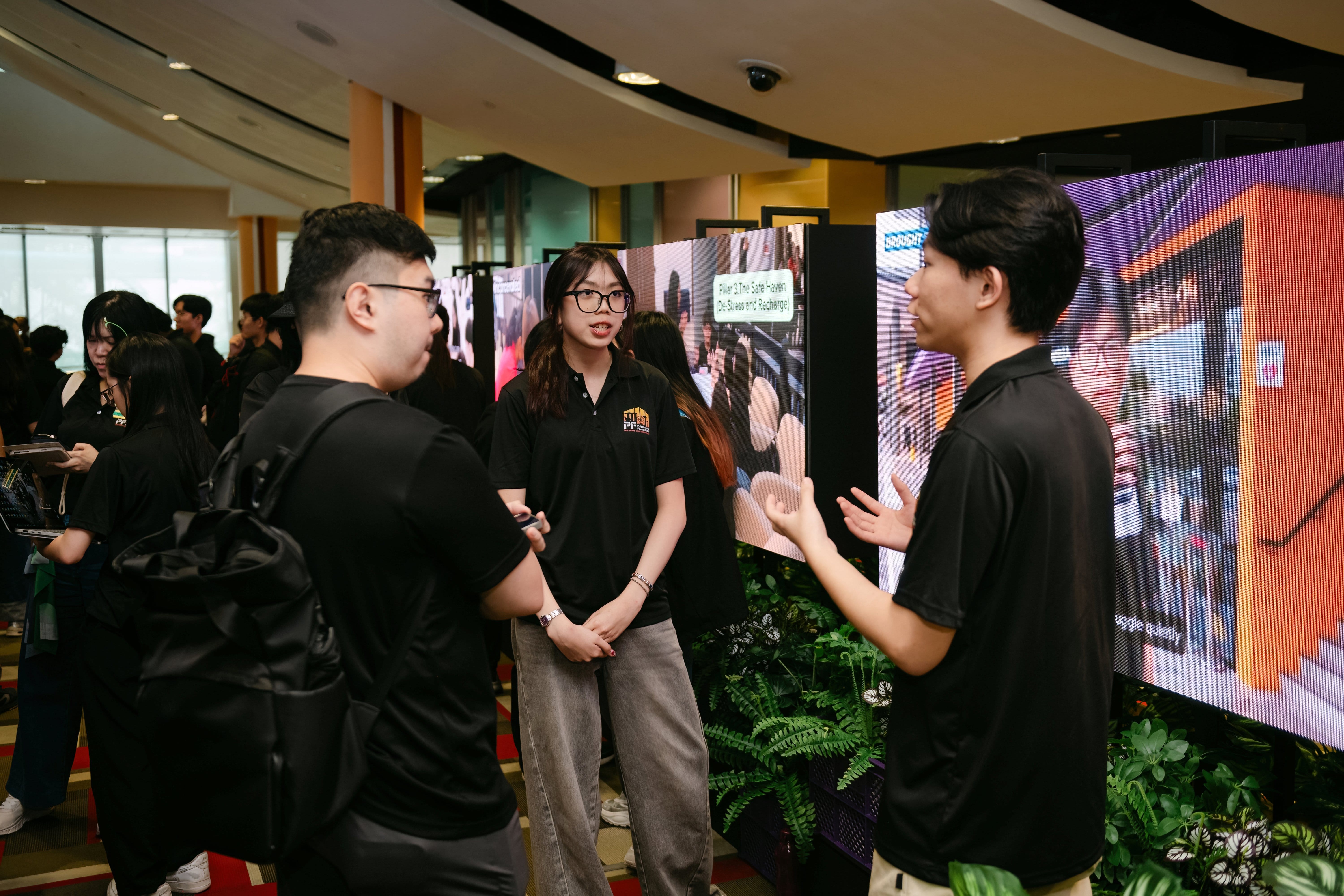 Three young people in black polo shirts converse at an indoor exhibition with display boards and a large screen.