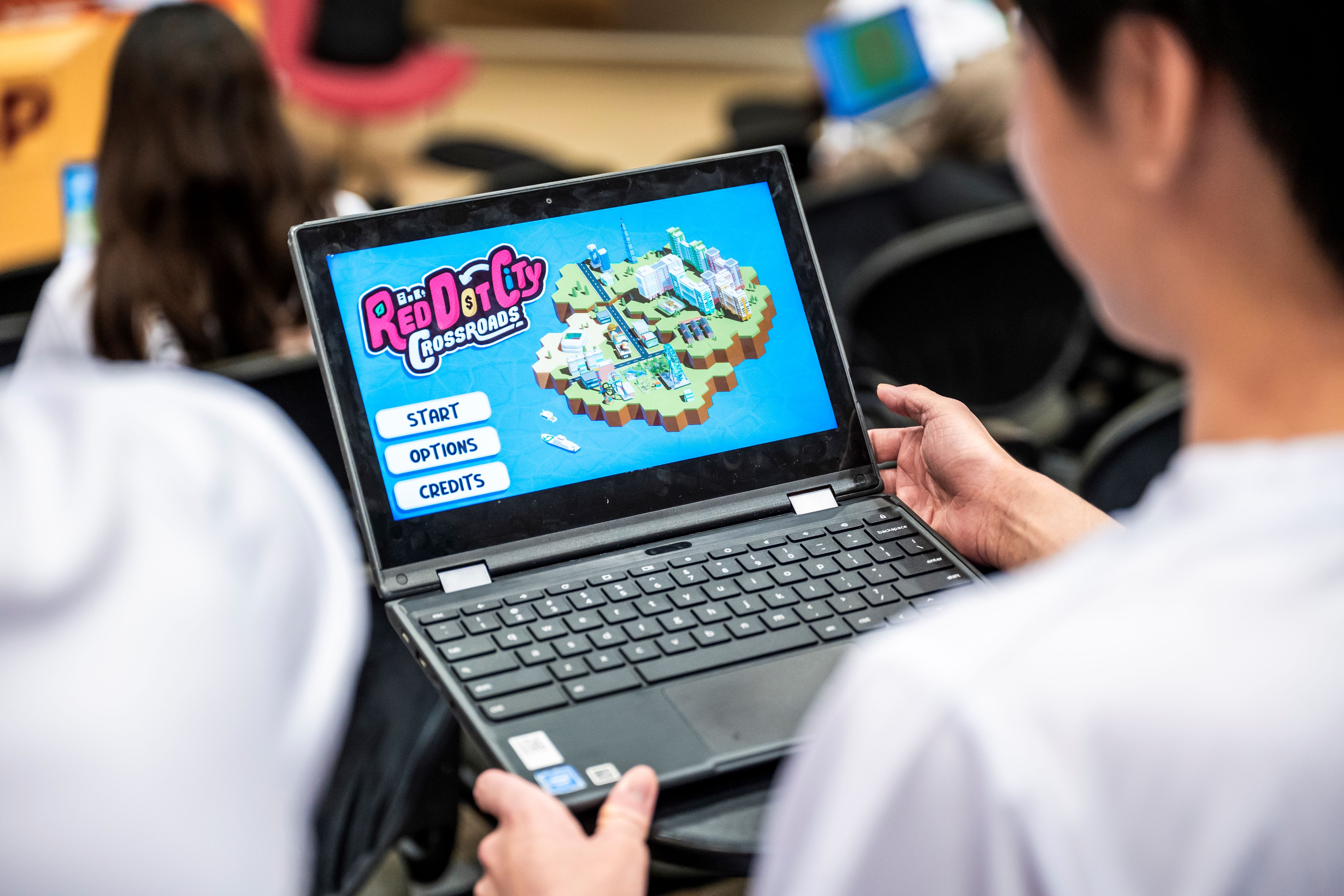 A student holds a laptop showing the Red Dot City Crossroads game menu with Start, Options, and Credits buttons.
