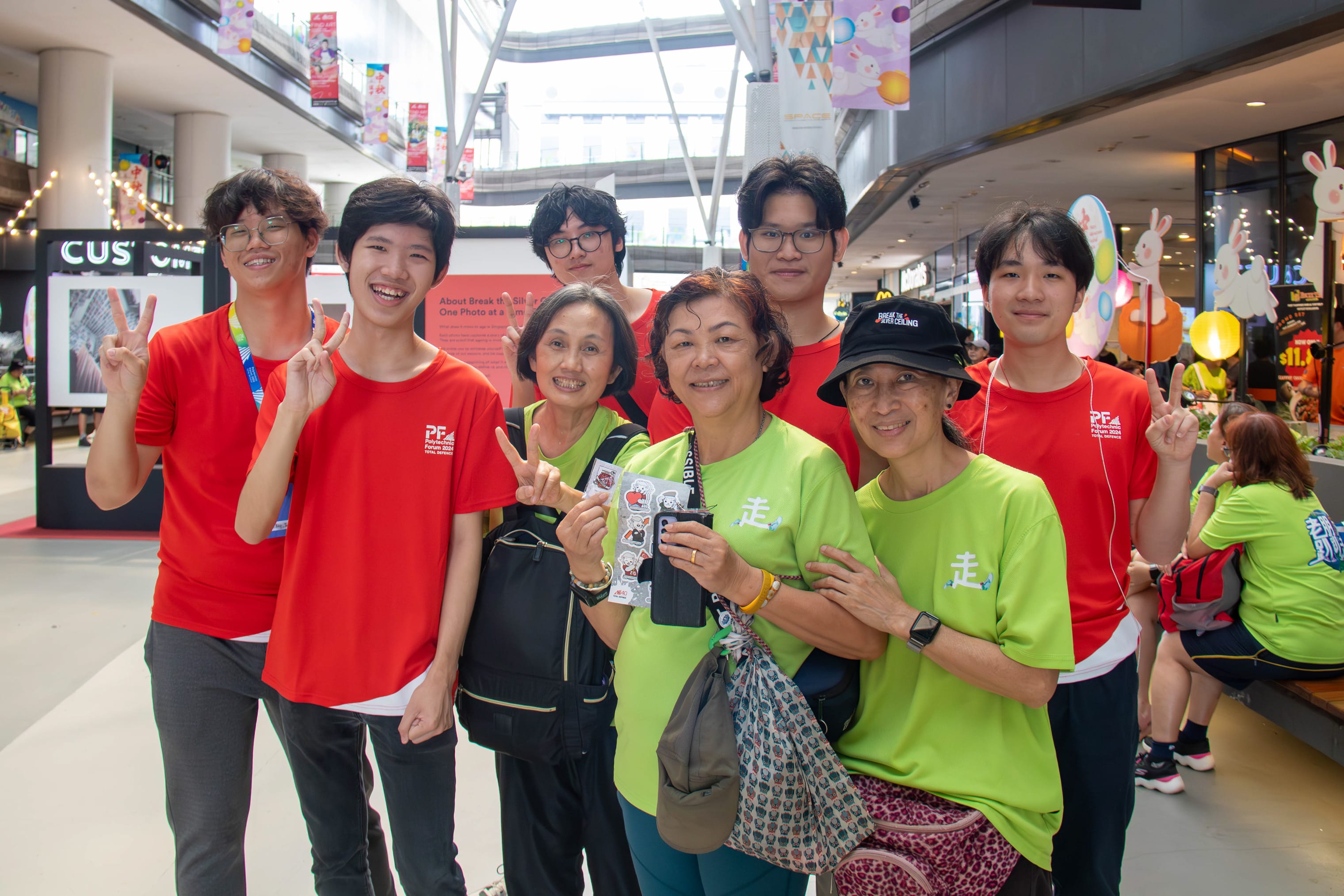 A group of eight smiling people in red and lime green shirts pose together indoors, some making peace signs.