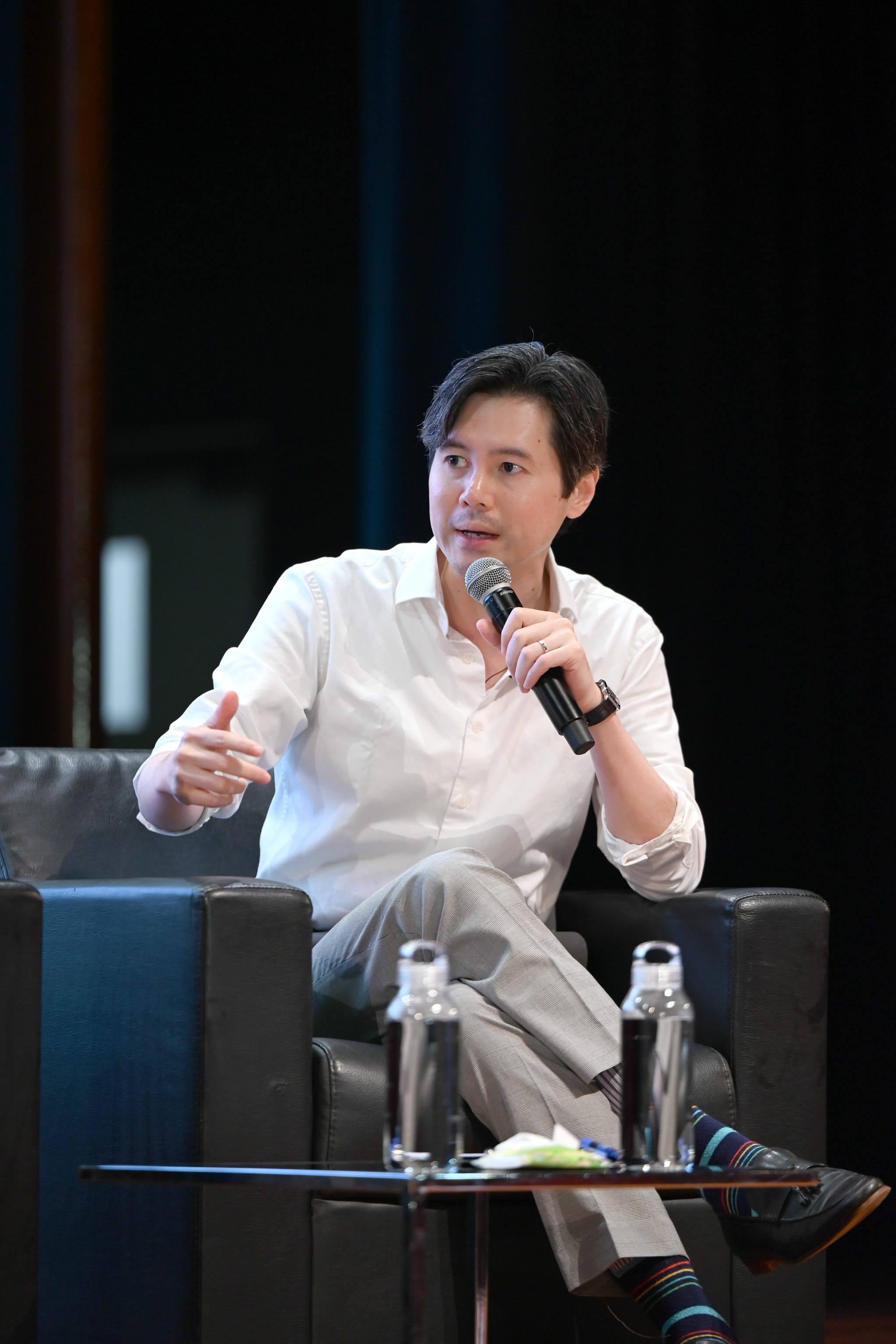 A man in a white shirt sits in a black armchair, holding a microphone and gesturing while speaking at an event.