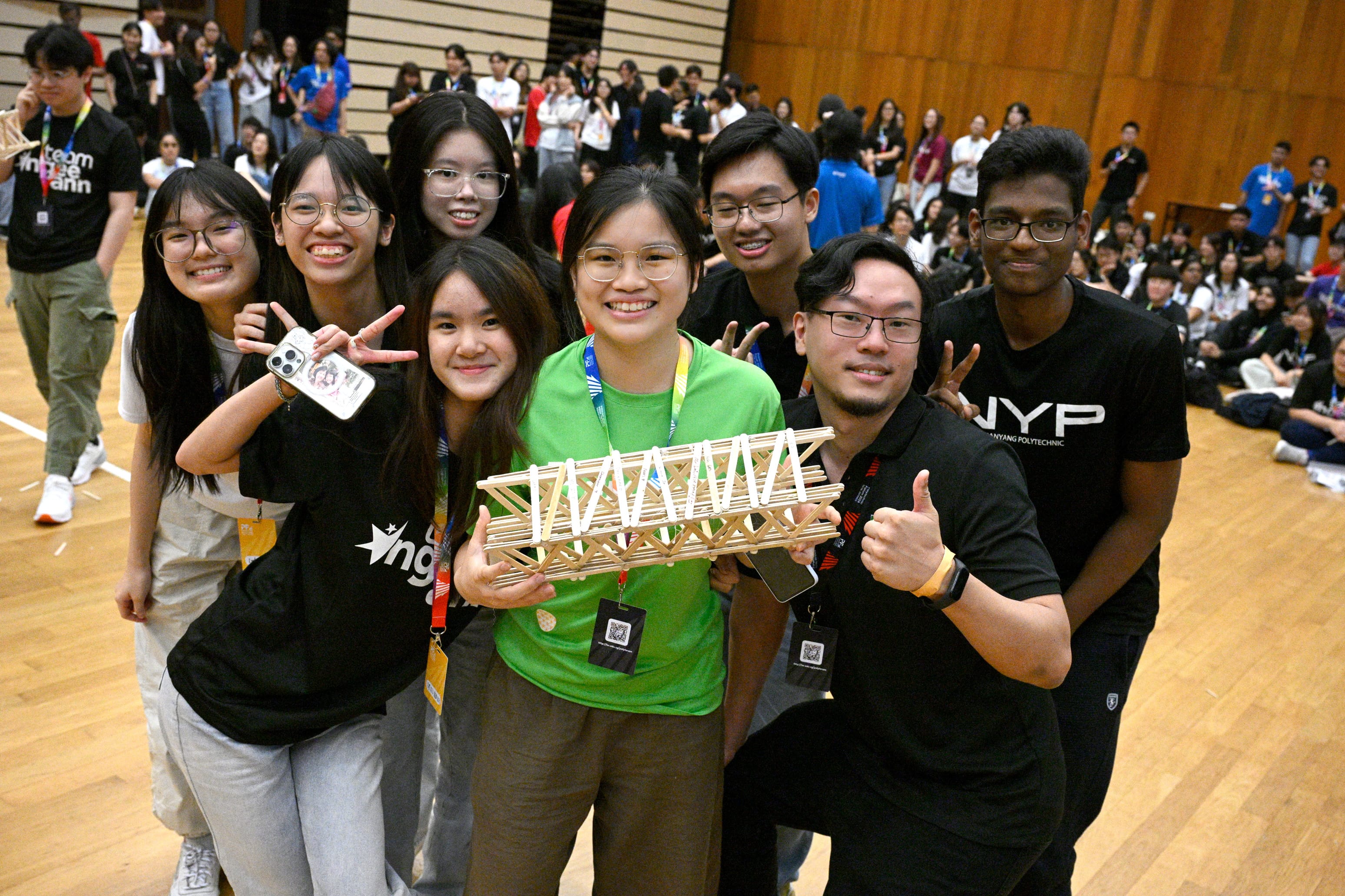 A group of smiling students poses together holding a small wooden bridge model in a large hall.