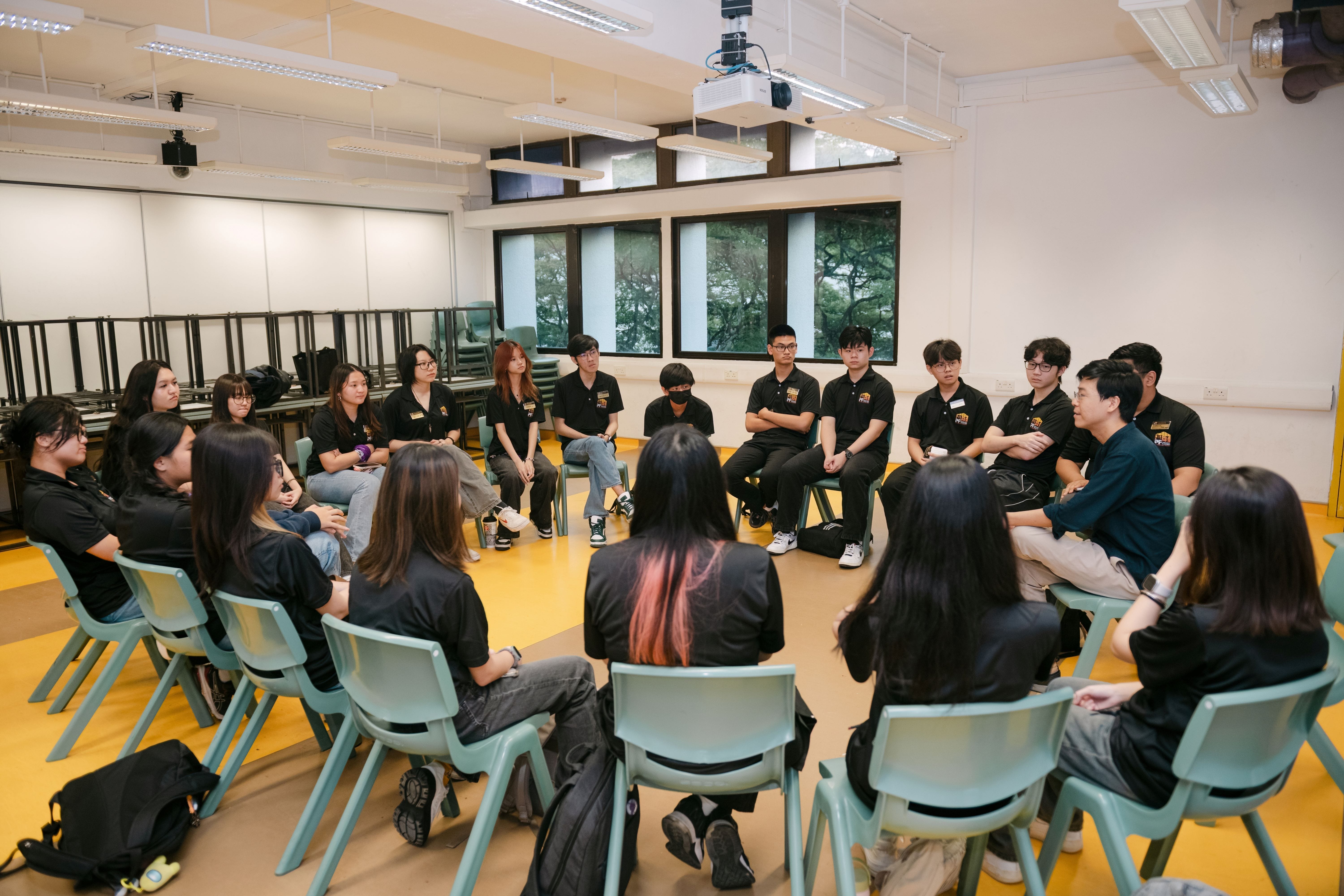 A group of about 20 young people in black shirts sit in a circle on light blue chairs in a bright classroom.