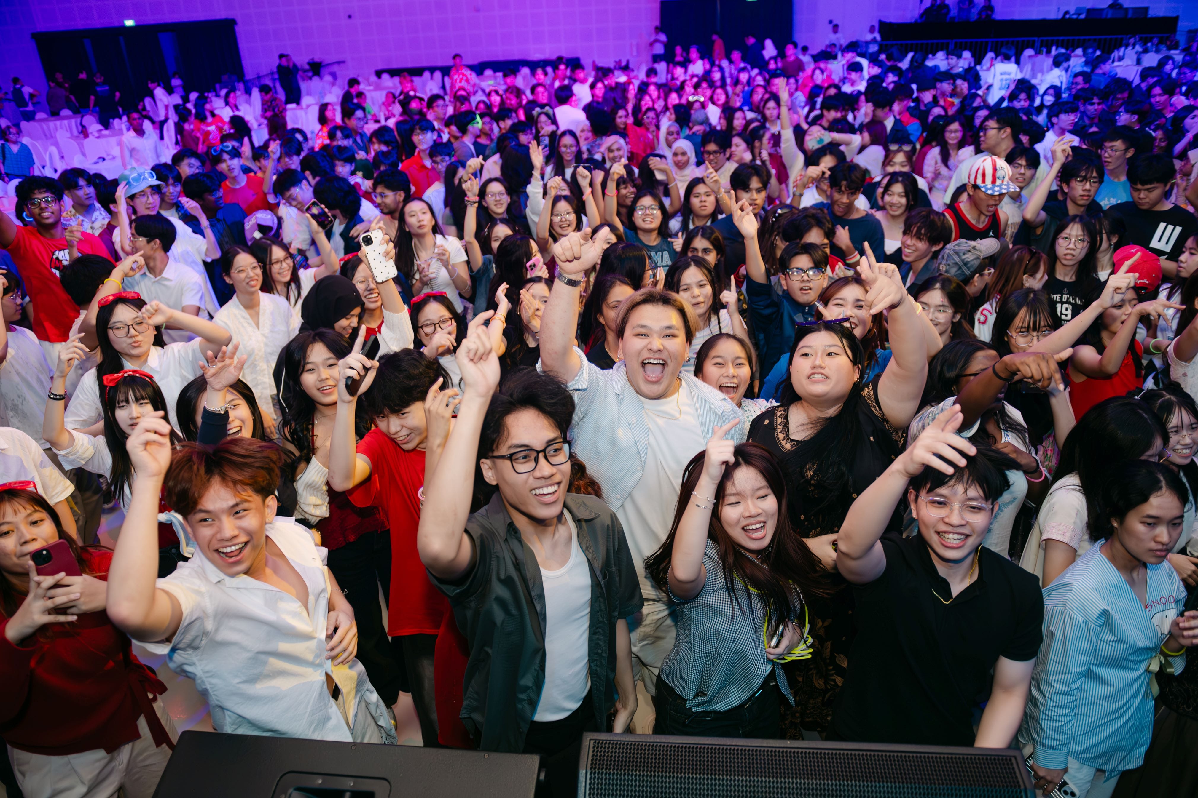 Large crowd of young people cheering with raised hands and big smiles at an indoor event with purple lighting.