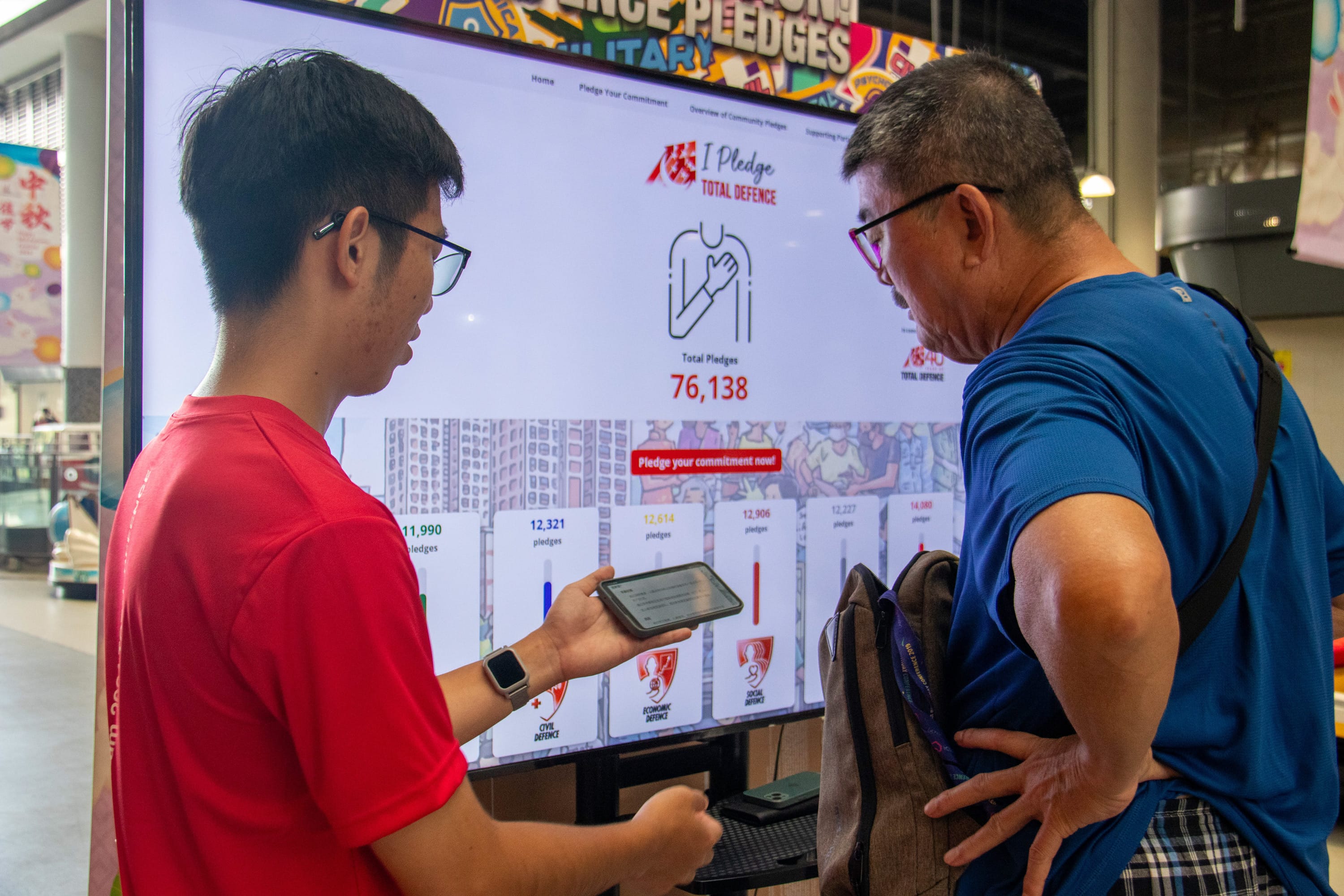 Two men look at a large screen showing an "I Pledge Total Defence" website with 76,138 total pledges.