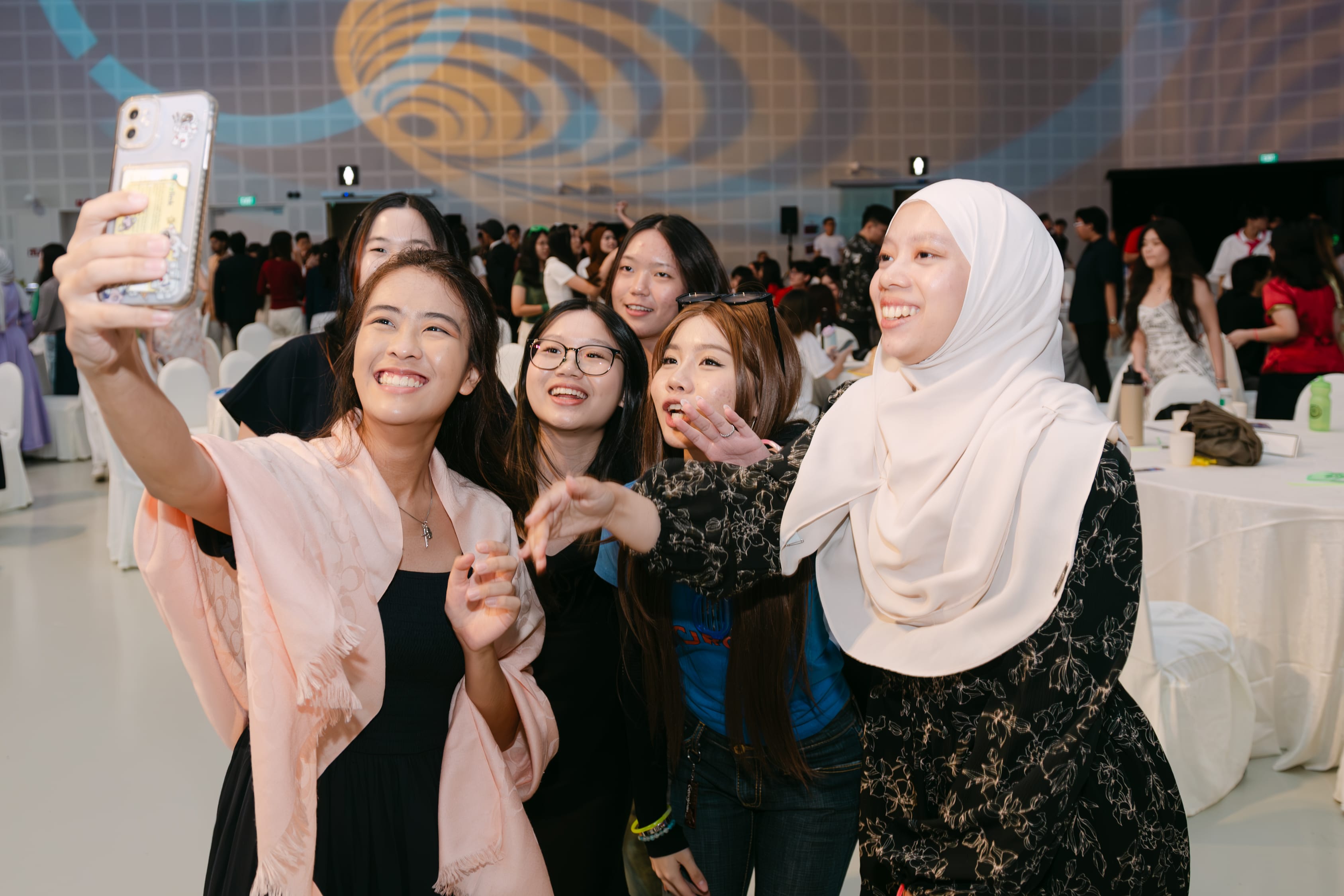 A group of smiling young women pose for a selfie at a crowded indoor event with a decorated backdrop.