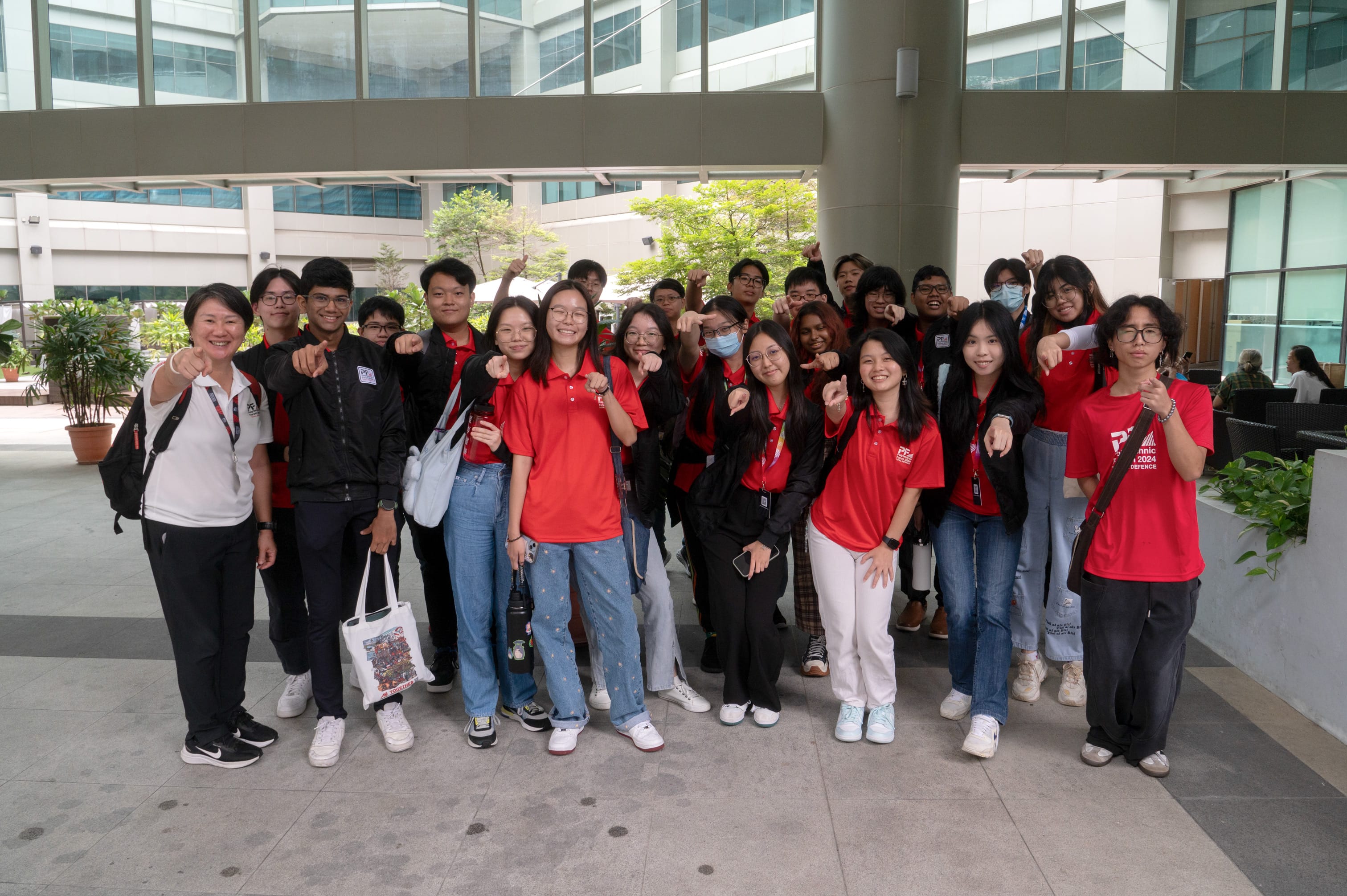 A large group of students in red and black shirts pose outdoors near a modern building, pointing at the camera.