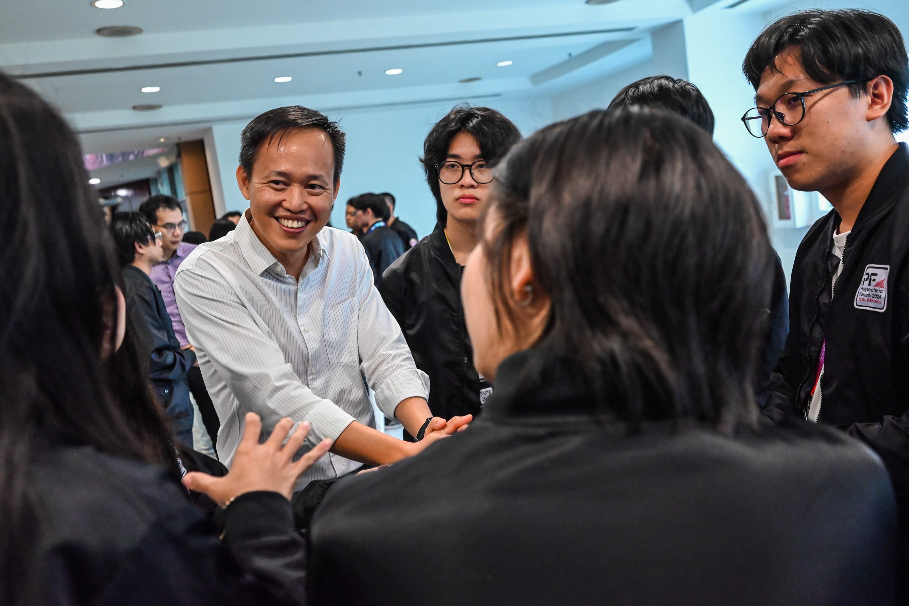 A smiling man in a white shirt engages in conversation with a group of young people in black jackets indoors.