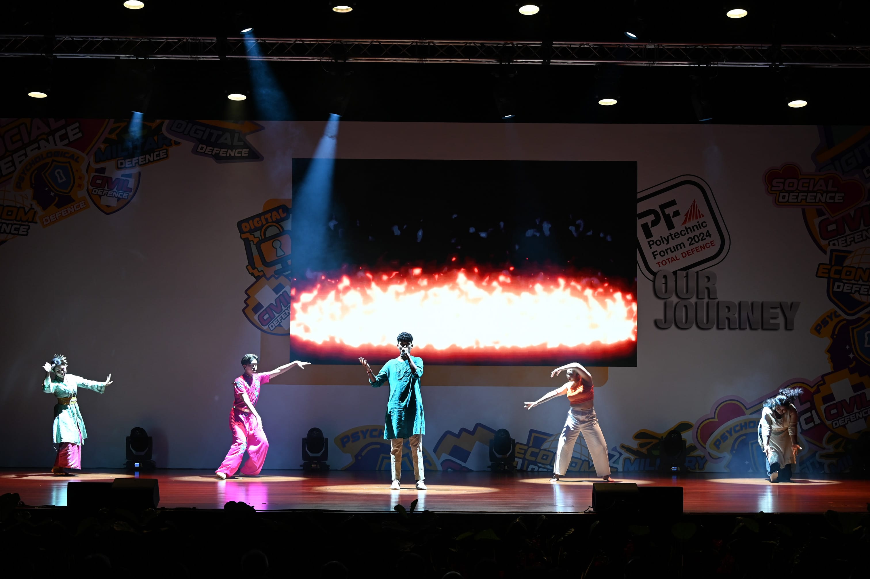 Five performers on a lit stage at Polytechnic Forum 2024, with a fire visual on screen and a Total Defence backdrop.
