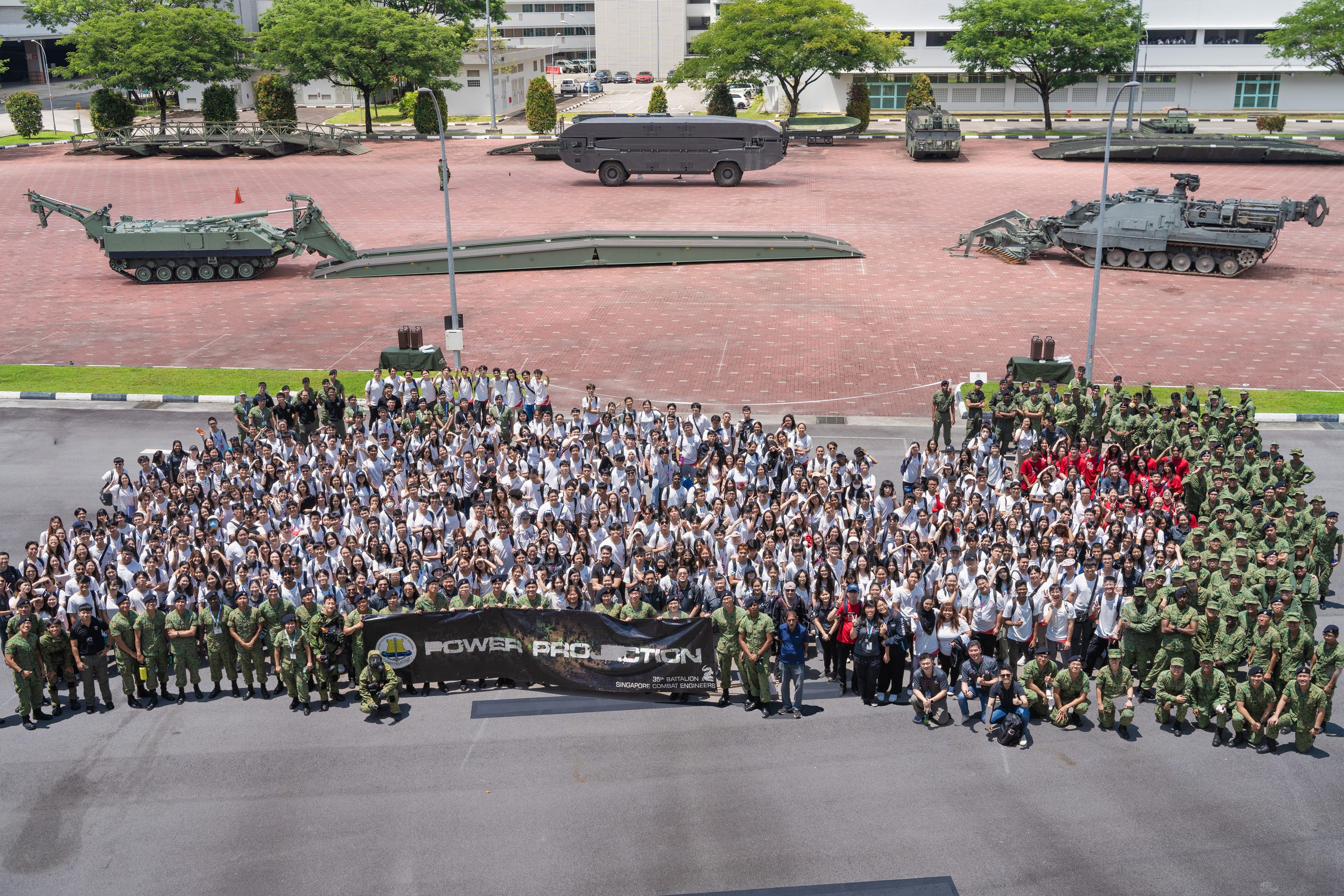 Large group of soldiers and civilians posing with a Power Projection banner in front of military vehicles.
