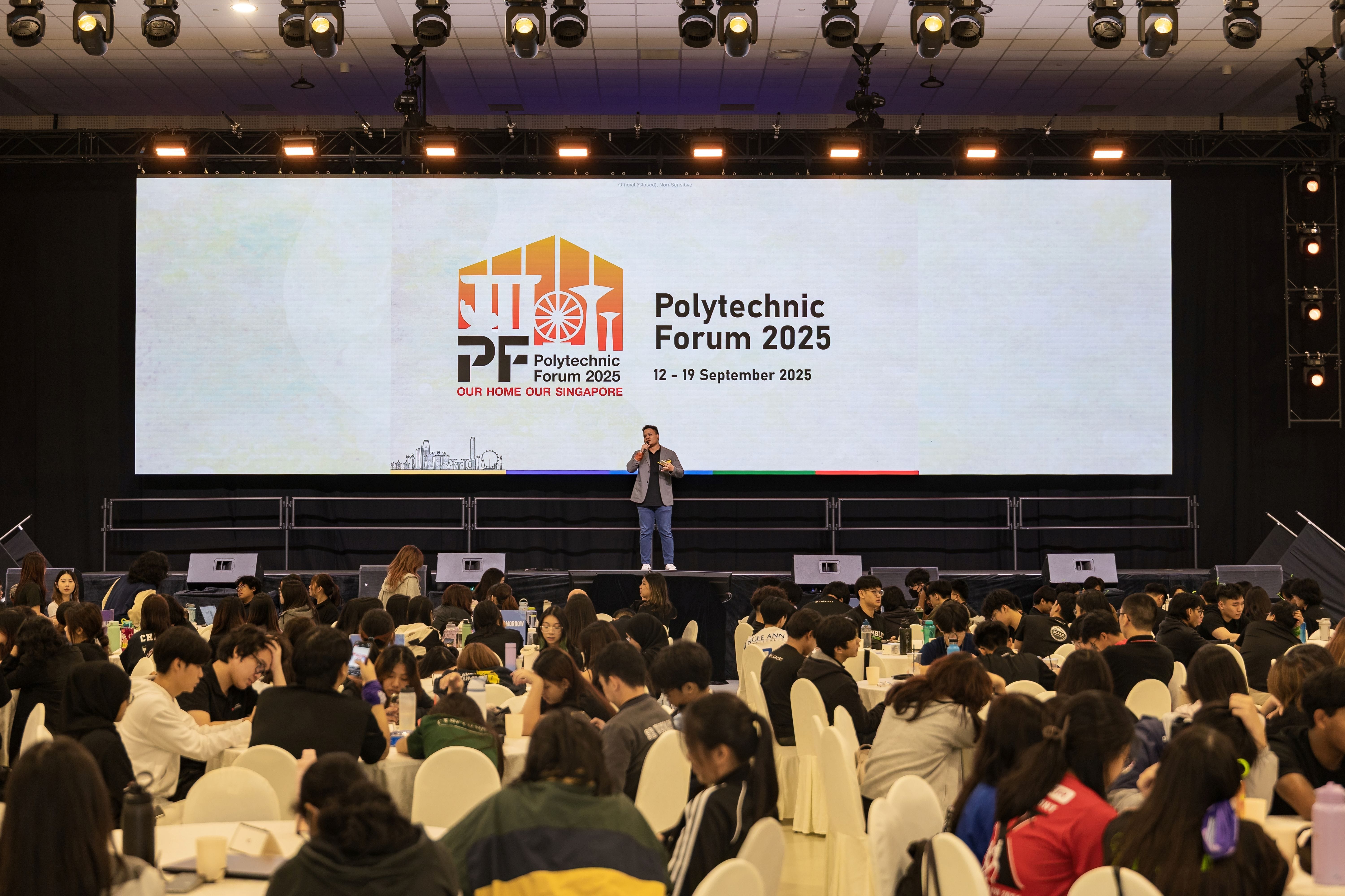 A speaker on stage at Polytechnic Forum 2025, 12-19 September, addressing a large seated audience in a hall.