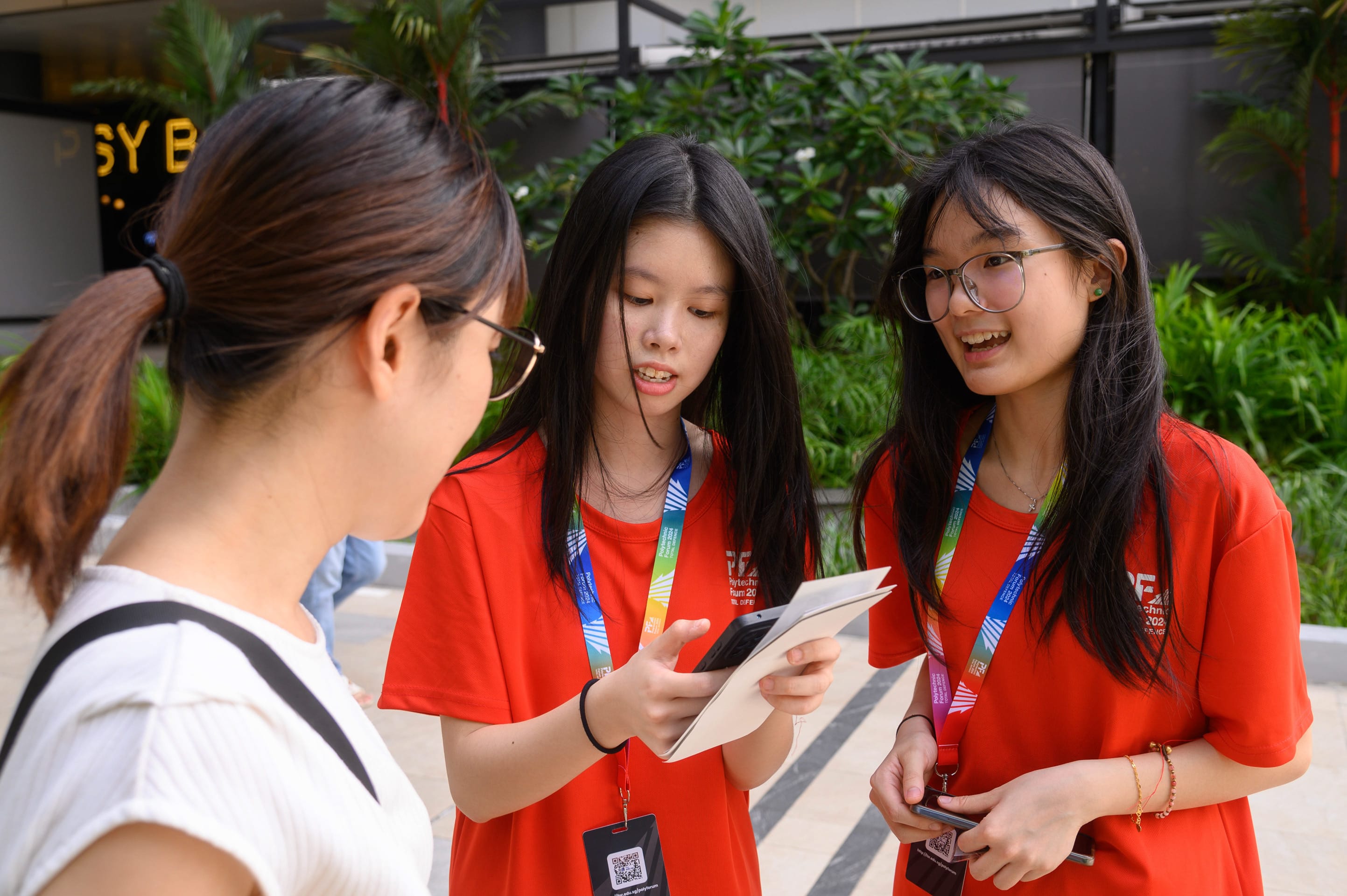 Two young women in red shirts with lanyards show a phone and booklet to another woman outdoors.