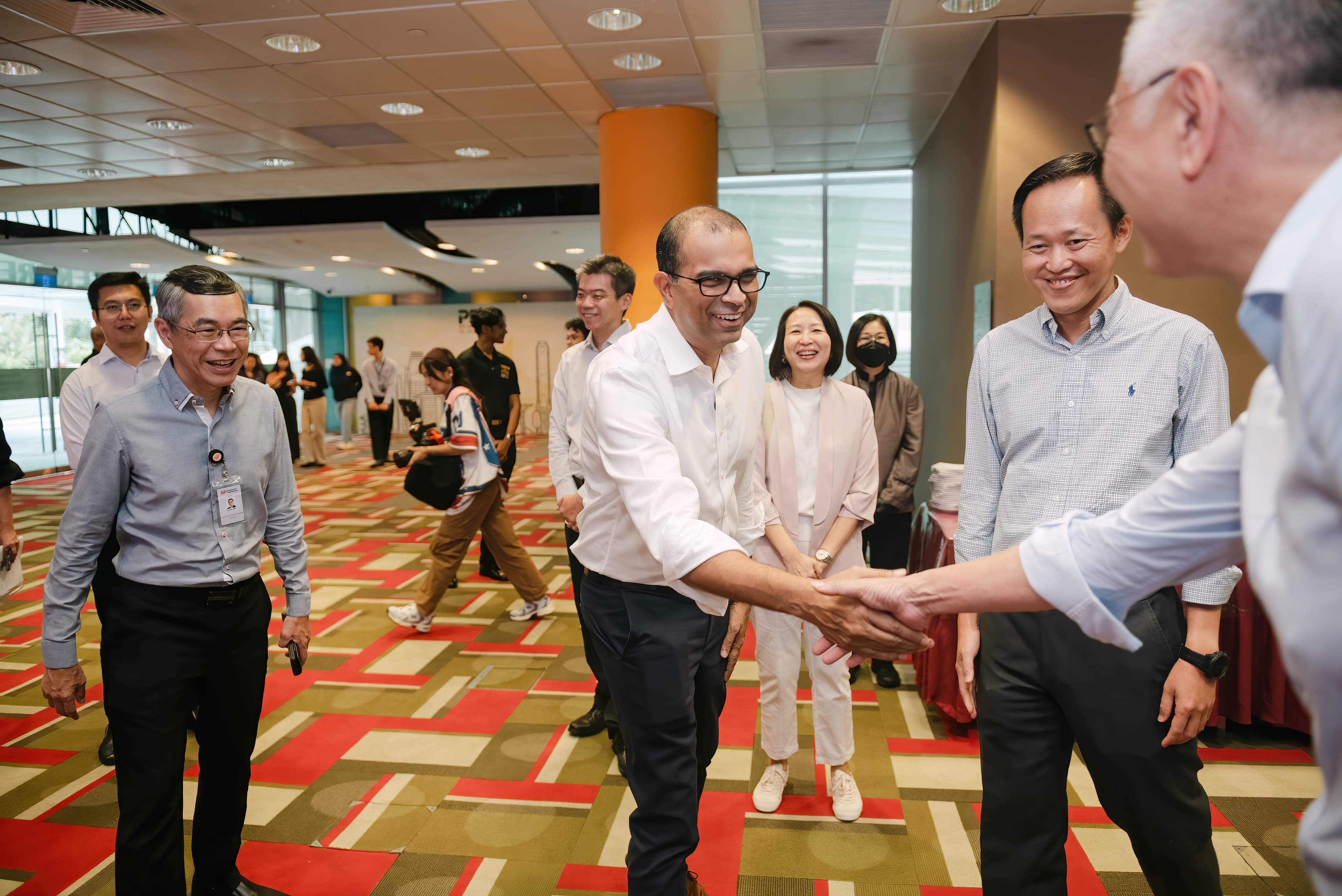 A man in a white shirt shakes hands with another man while smiling, surrounded by a group of people in a modern hall.