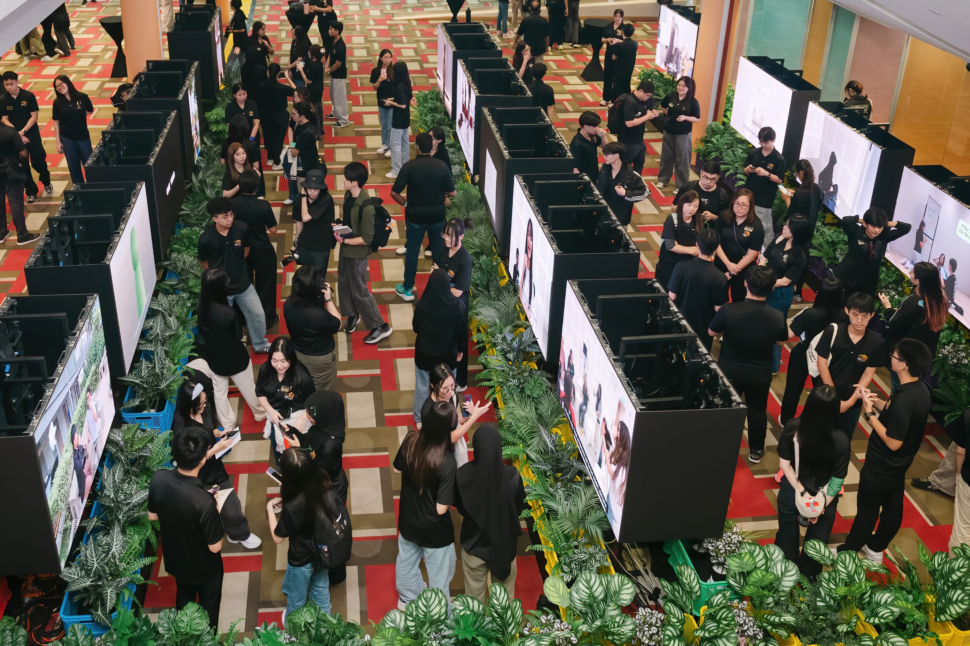 Large group of people in black shirts gathered around rows of display screens and plants in a decorated hall.