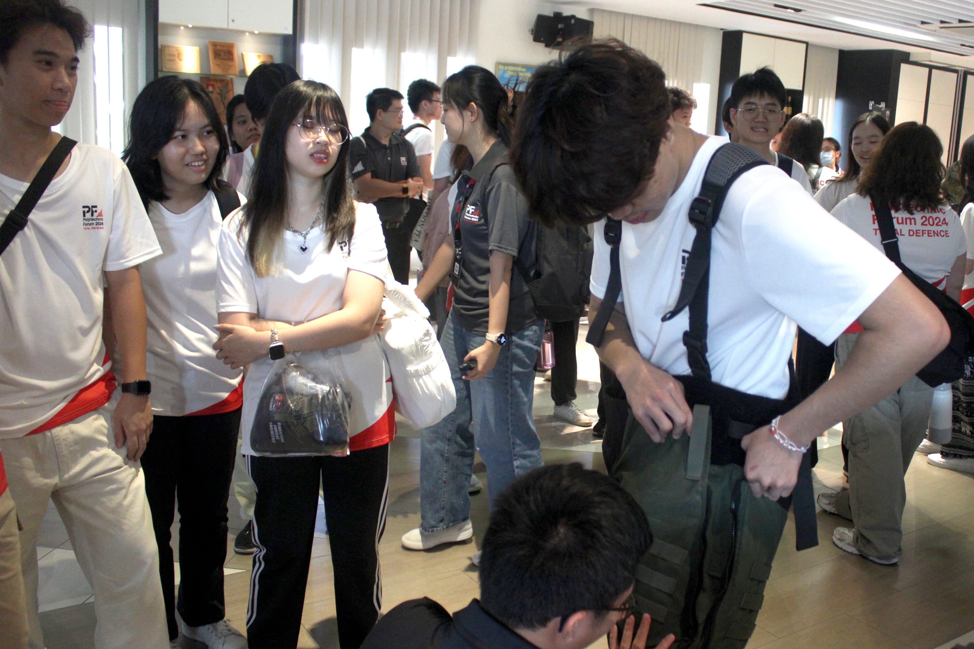 A group of young students in white Polytechnic Forum 2024 shirts gather and chat indoors.