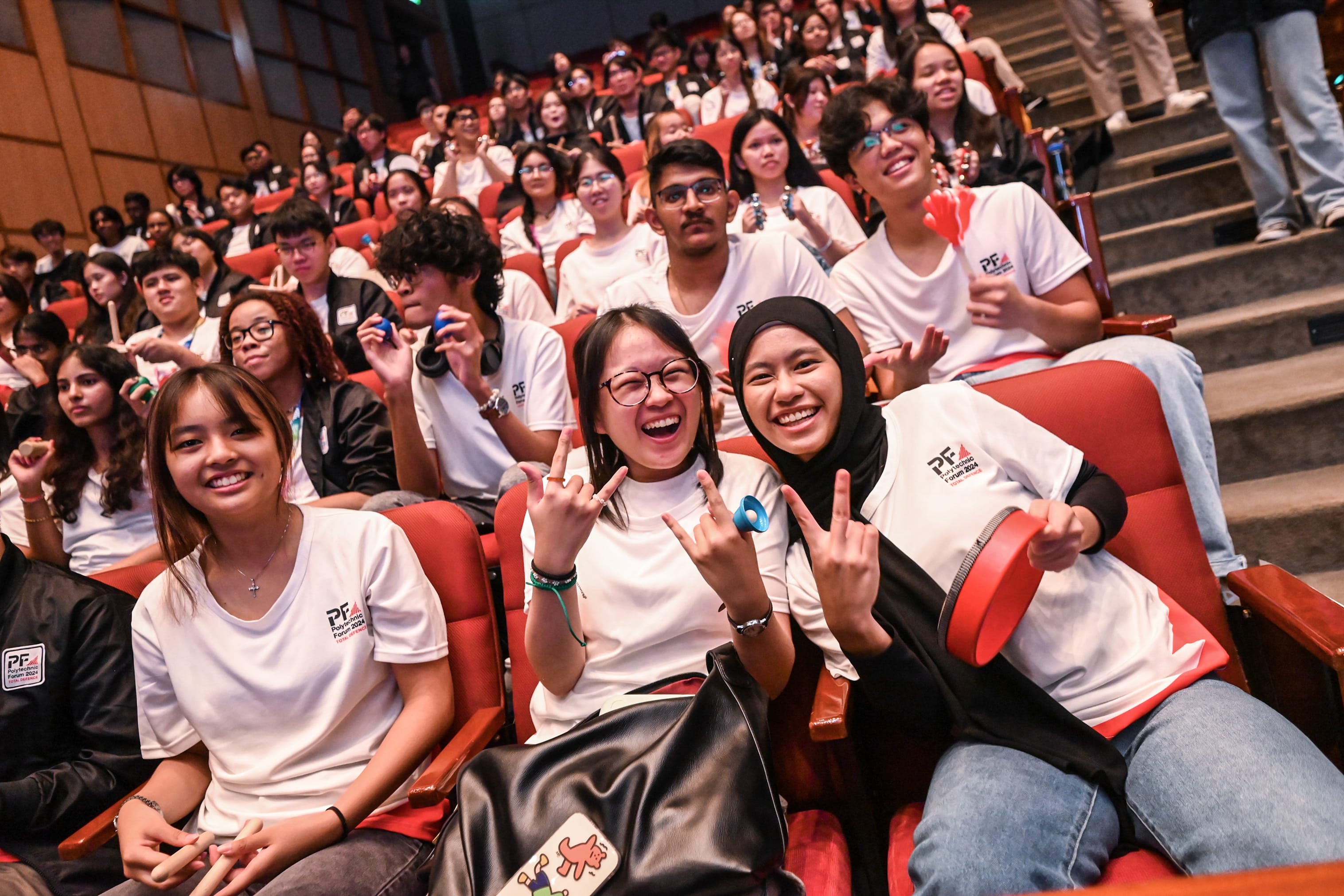 A large group of smiling young people in white PF Forum 2024 shirts sit in red auditorium seats, some making hand gestures.