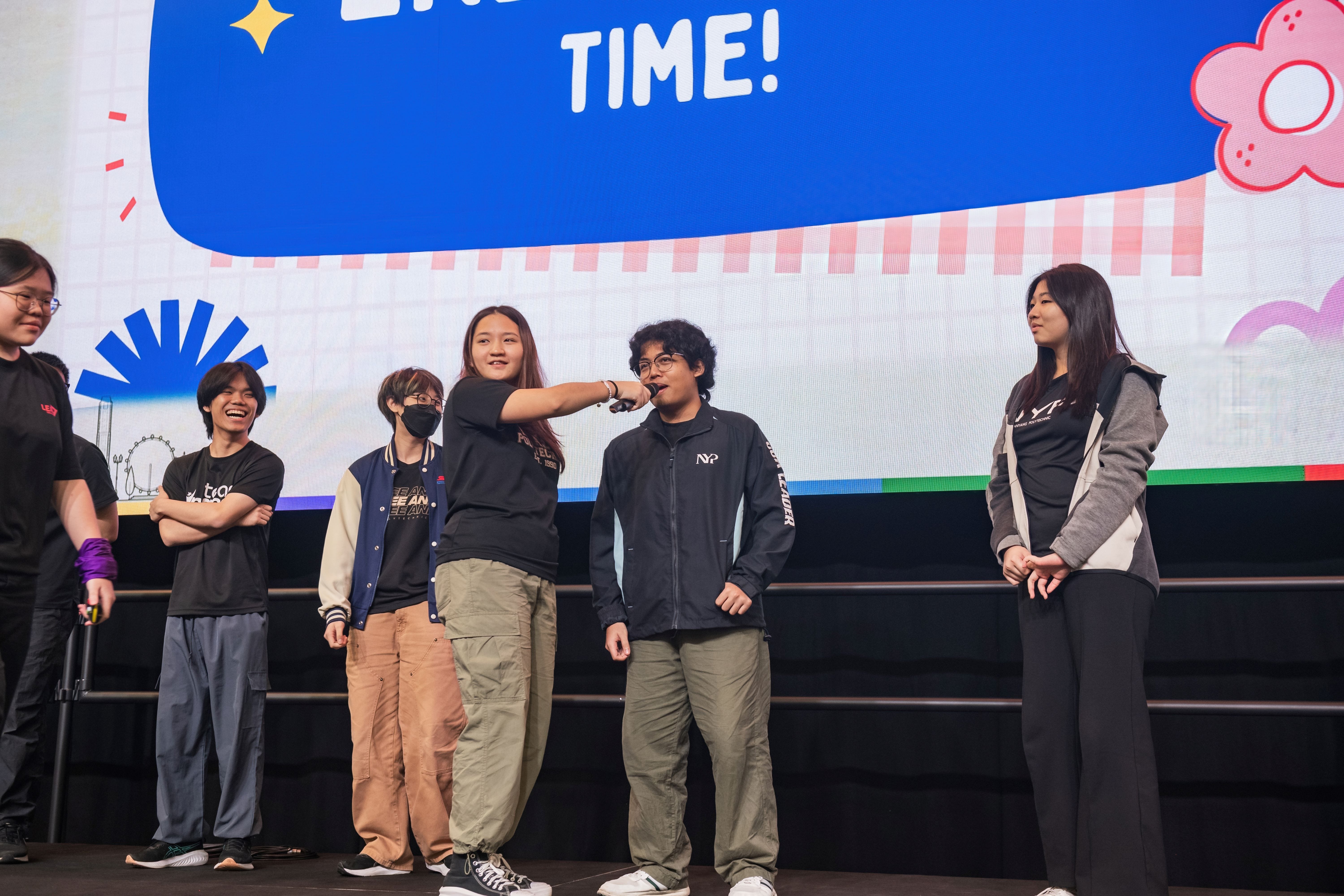 Six young people stand on a stage in front of a blue screen displaying the word TIME!