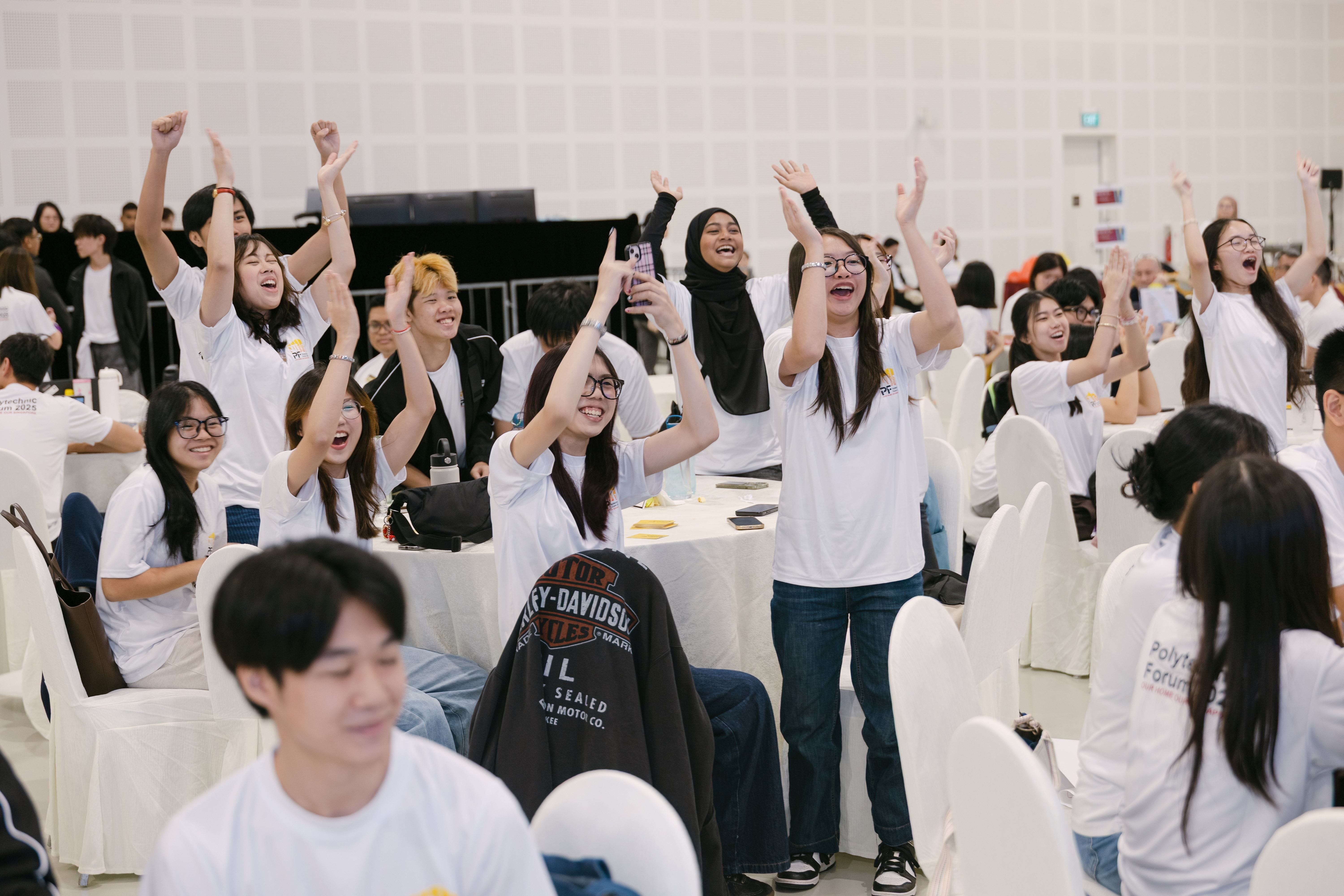 A crowd of young people in white t-shirts cheer with raised arms at a large indoor event with round tables.