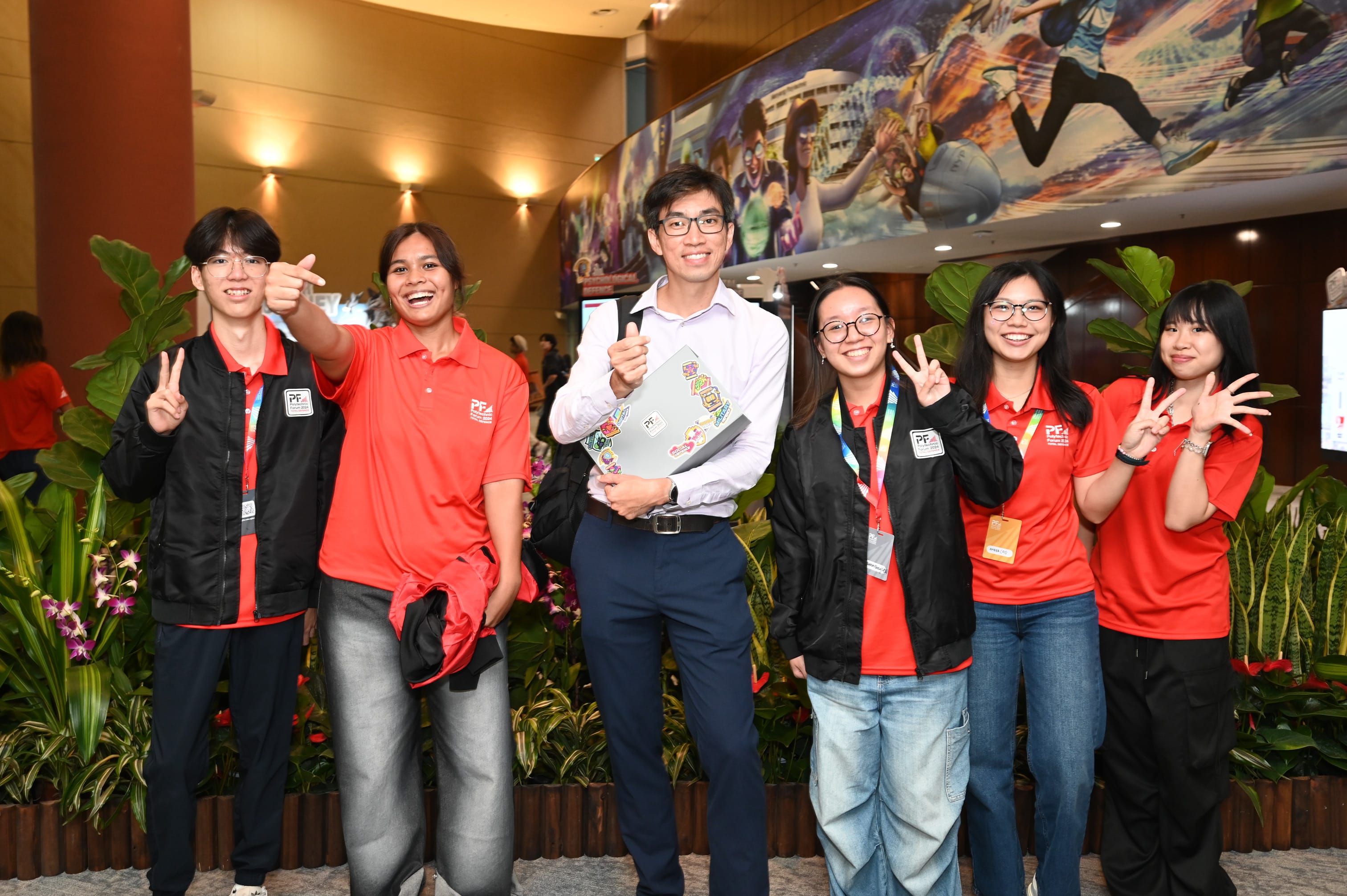 Six smiling young people in red shirts pose indoors, making peace signs, with a colorful mural behind them.