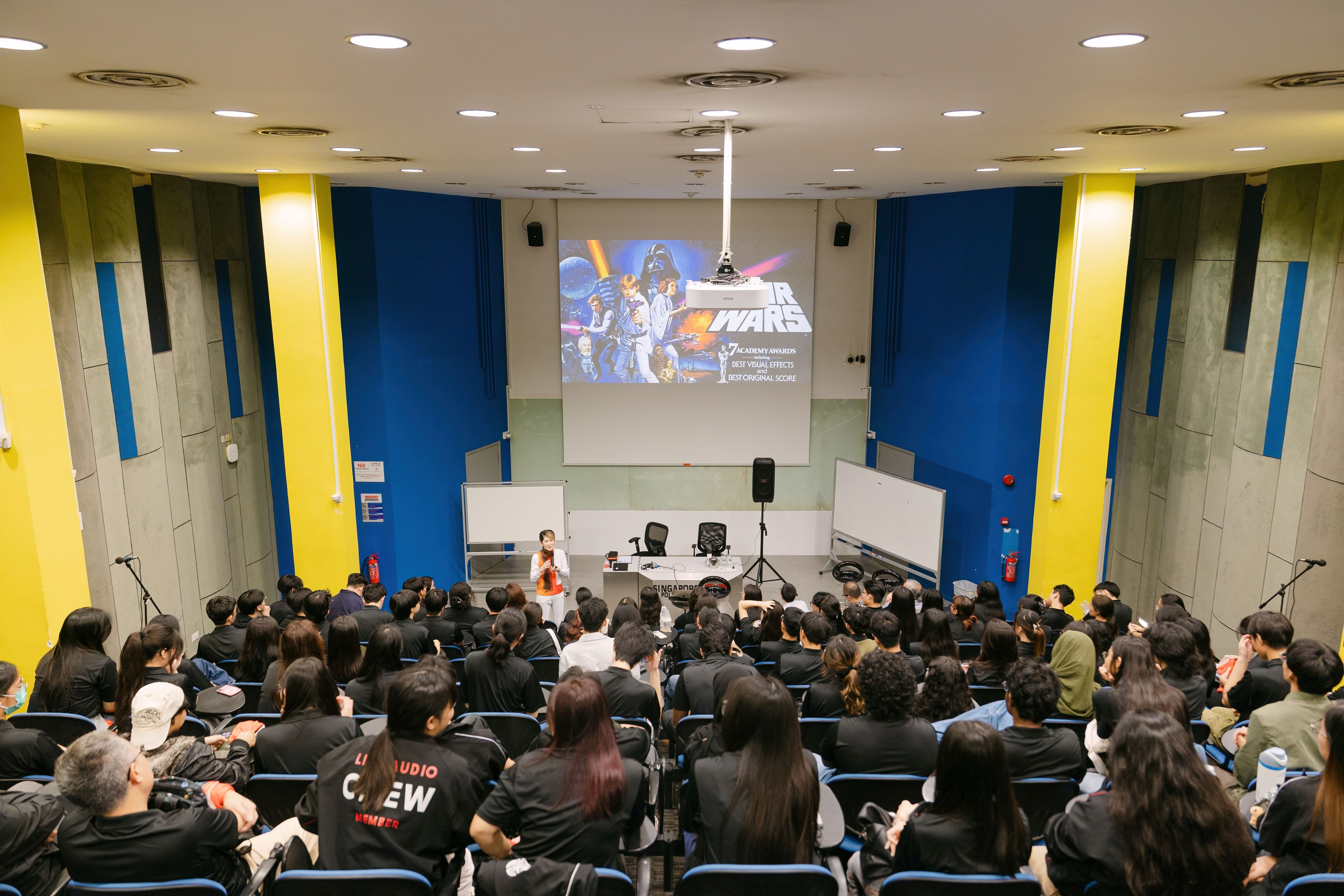 A packed lecture hall with a Star Wars movie poster projected on screen, audience seated facing a presenter at the front.
