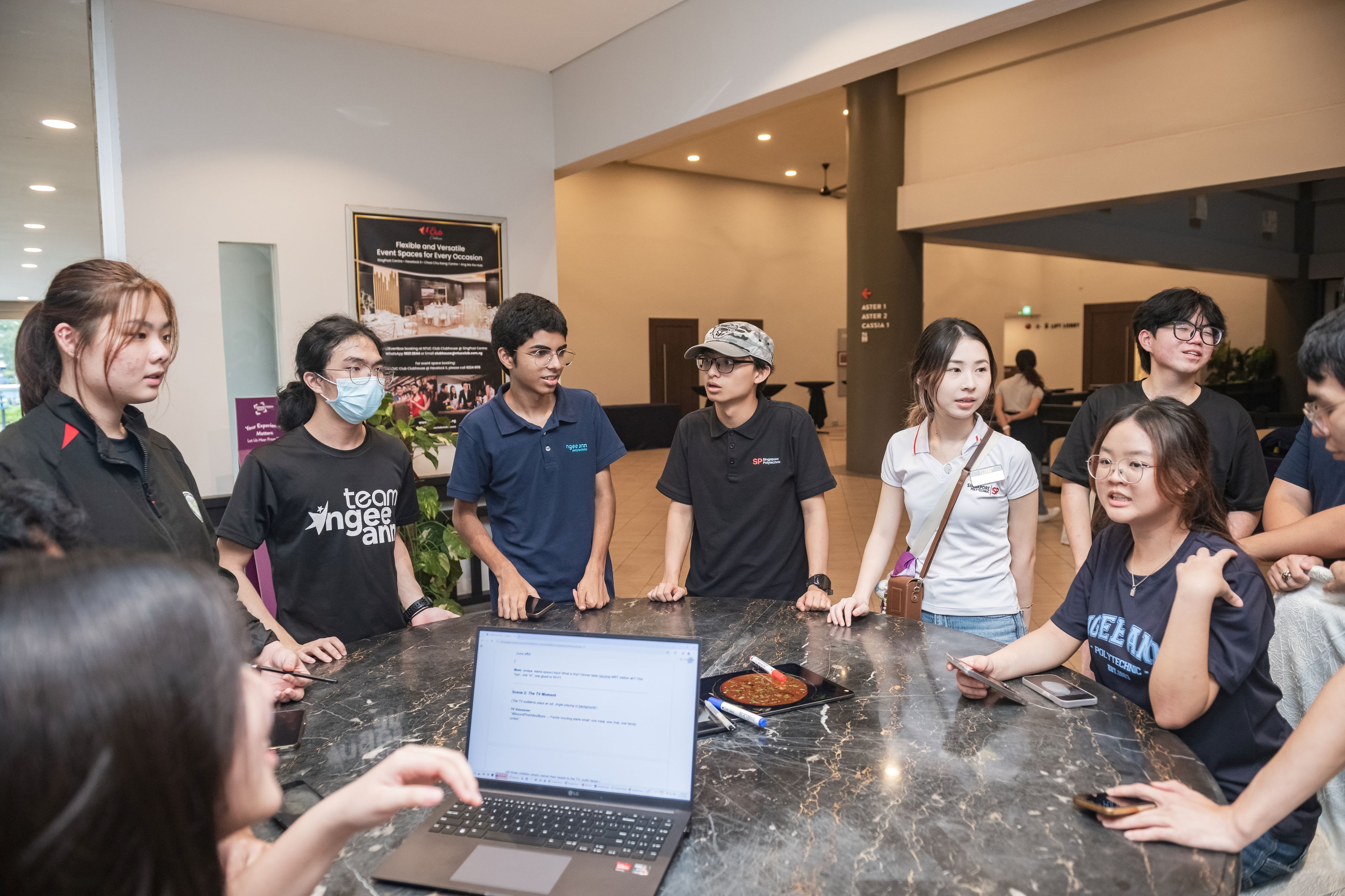 A group of young students gathered around a marble table with a laptop, engaged in discussion indoors.