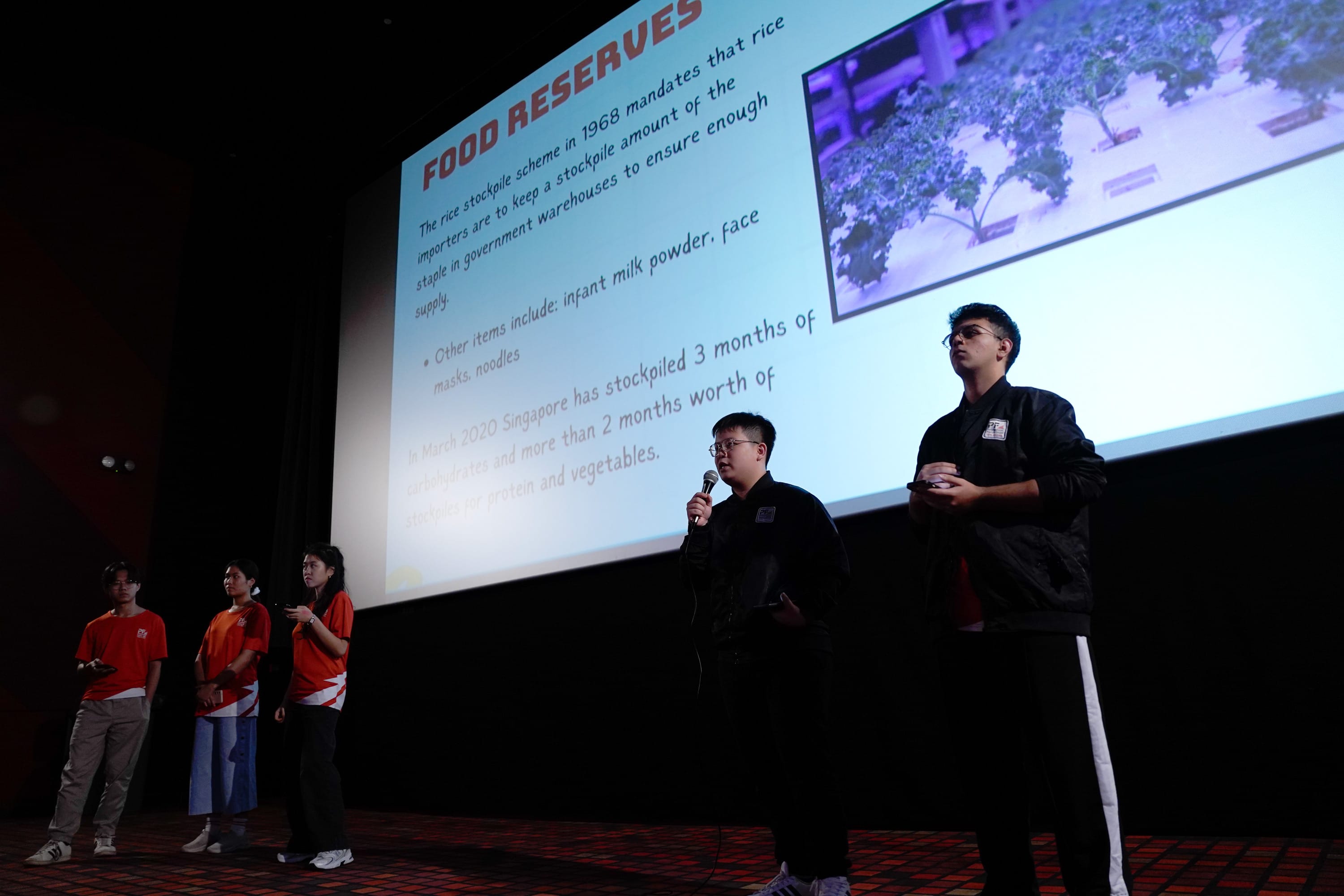 Five students present on stage before a large slide titled Food Reserves with text about Singapore's stockpile scheme.