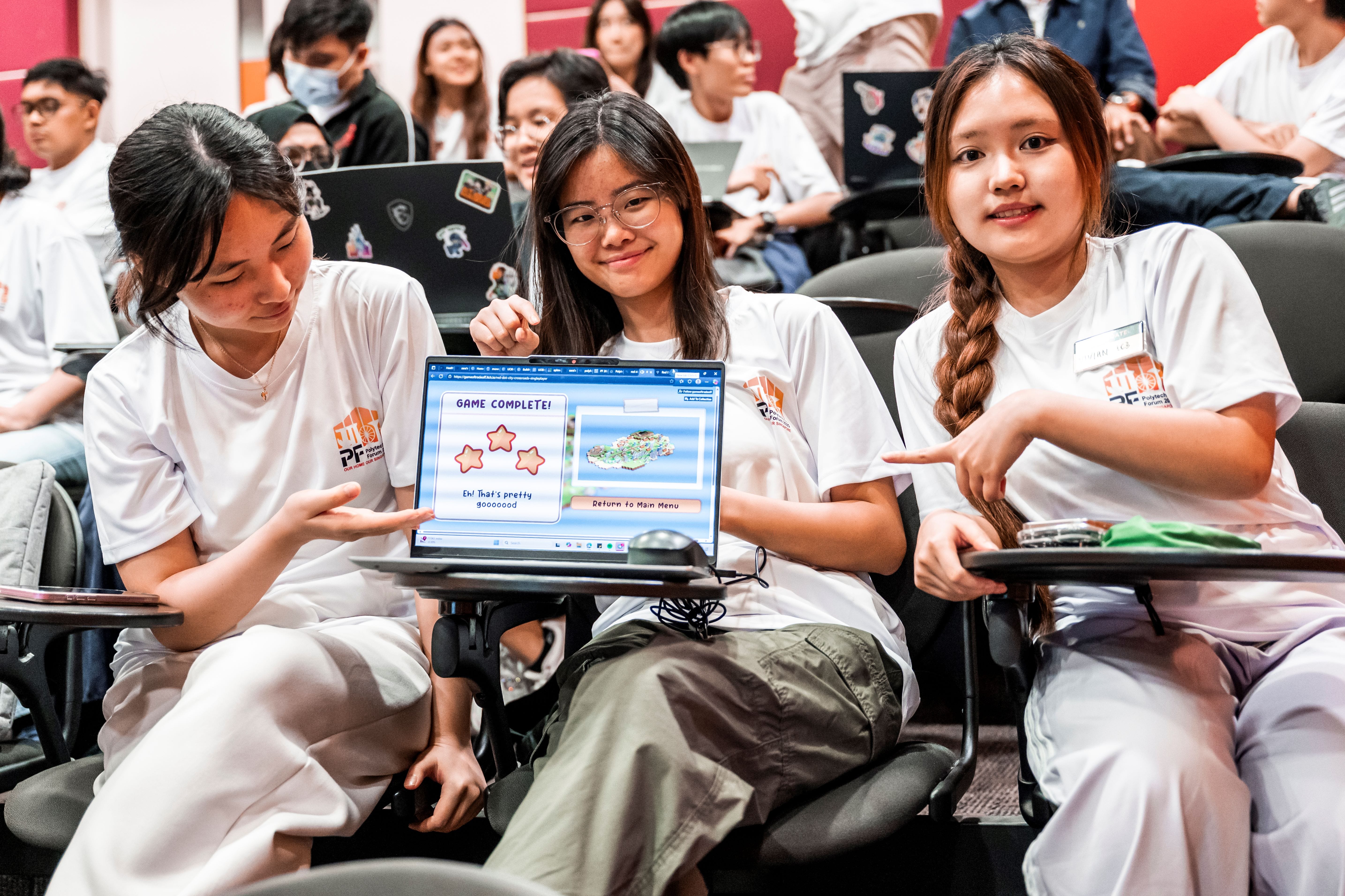 Three young women in white shirts smile and point at a laptop showing a "Game Complete" screen in a lecture hall.