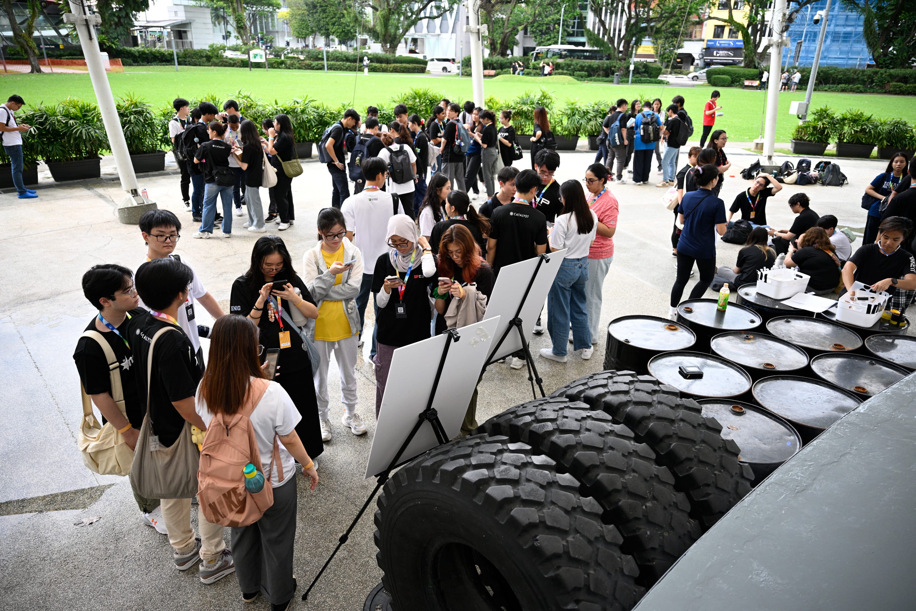 Large group of young people with lanyards gathering outdoors near a large vehicle tire, white boards, and metal barrels.