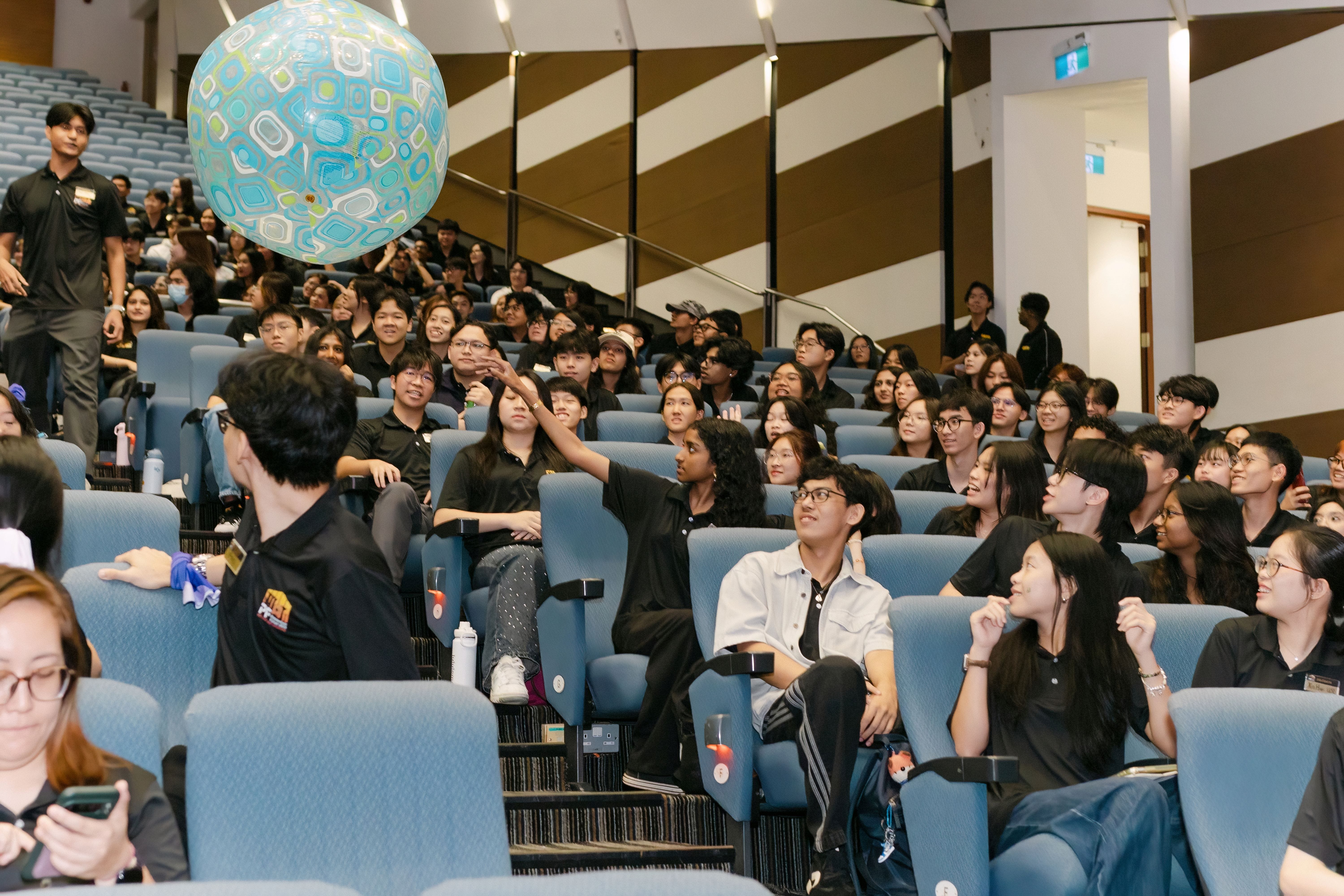 Large group of students in black uniforms seated in a tiered auditorium, watching a colorful inflatable ball float above them.