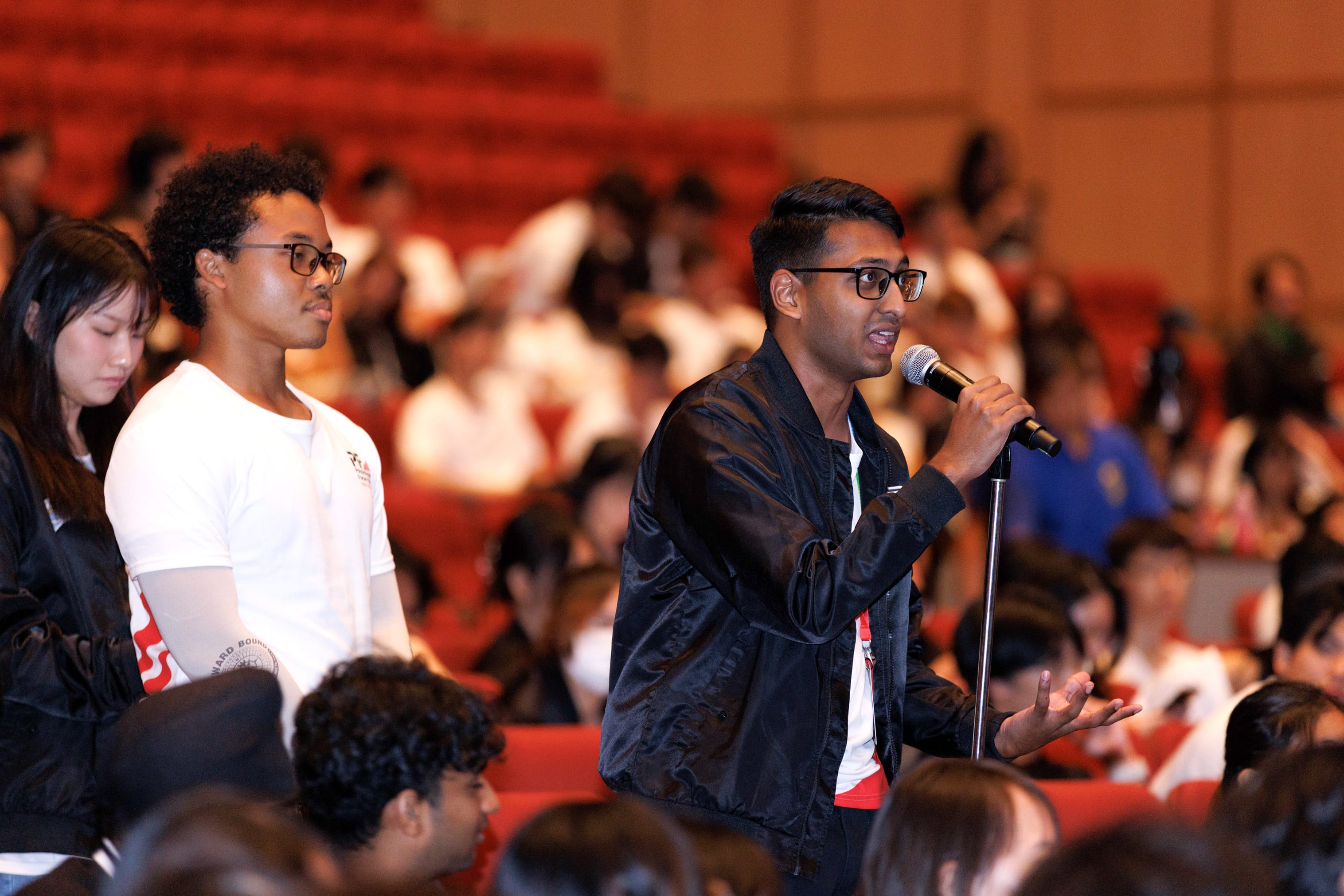 A young man in a black jacket speaks into a microphone in an auditorium filled with seated attendees.