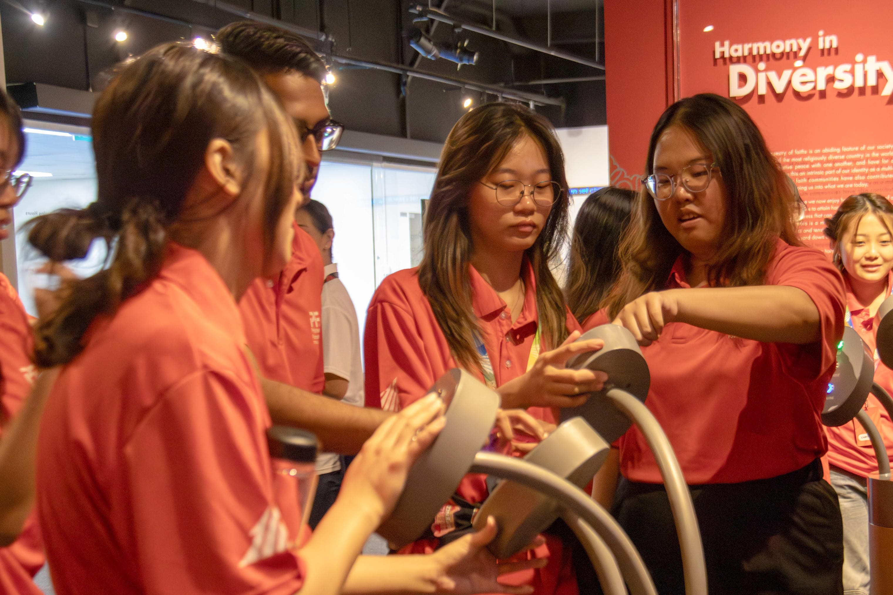Students in red uniforms examine an interactive exhibit near a red wall reading Harmony in Diversity.