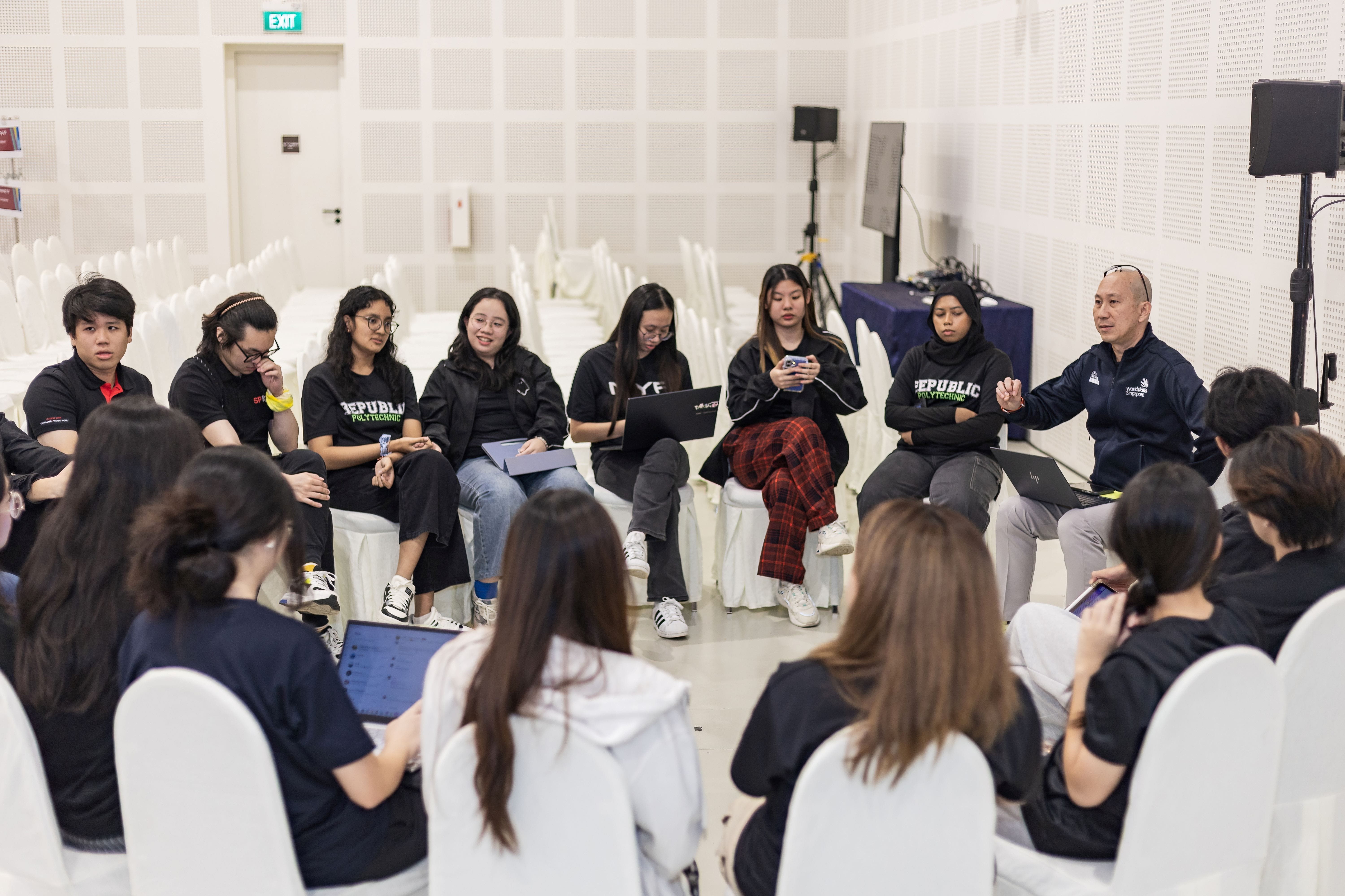 A group of Republic Polytechnic students and an instructor sit in a circle during a discussion session indoors.