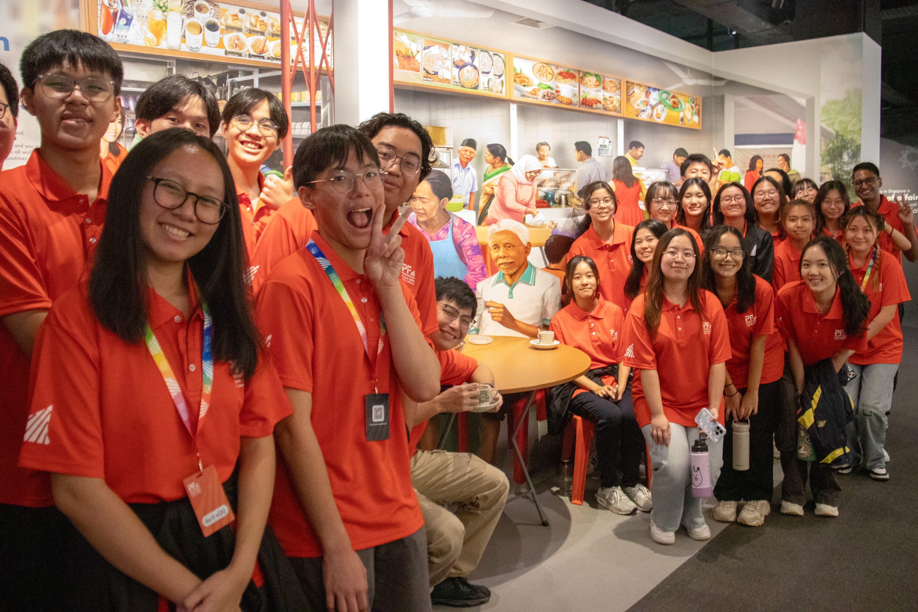 A large group of young people in red polo shirts pose smiling in front of a colorful food court mural display.