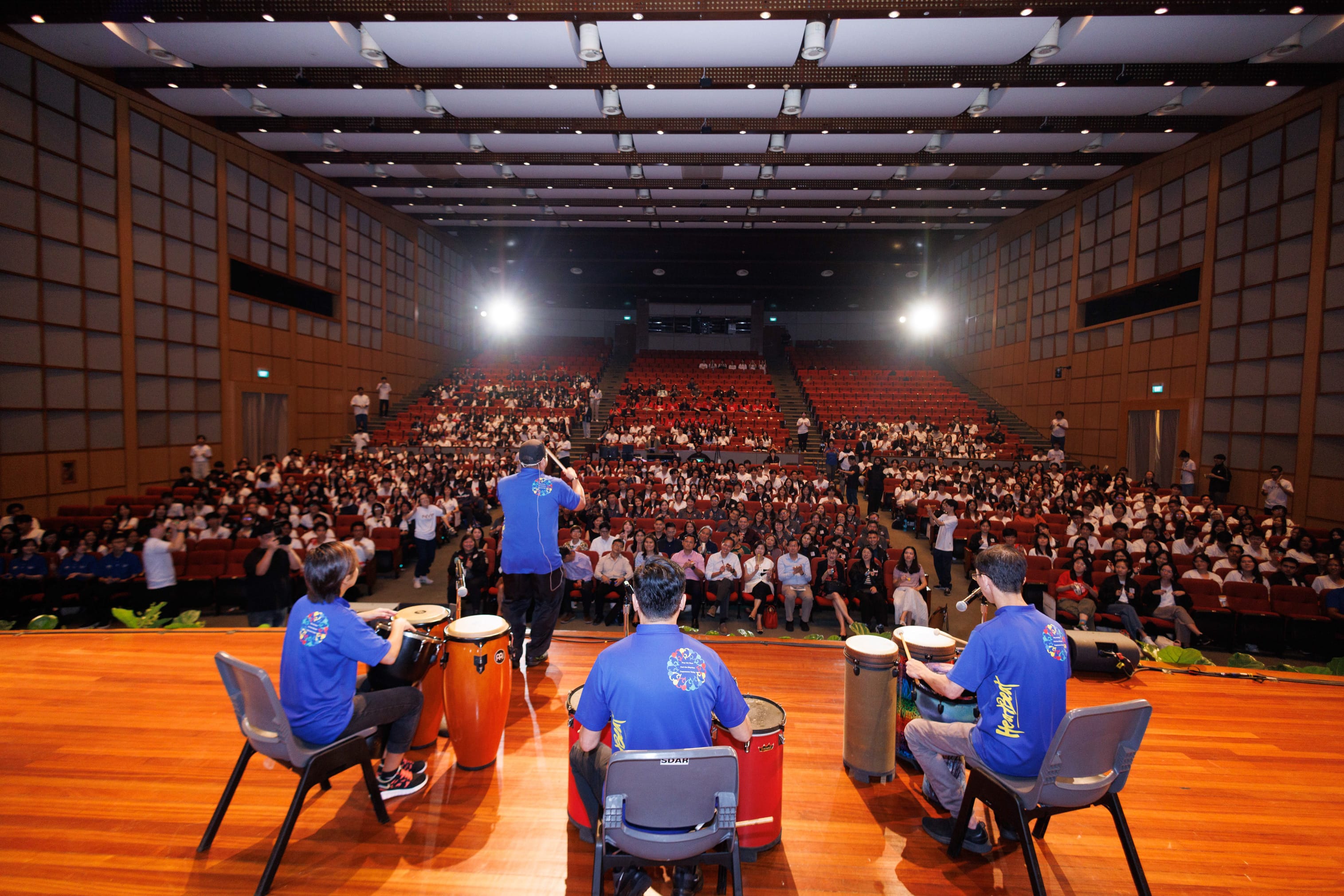 Four performers in blue shirts play drums on a wooden stage before a large seated audience in a concert hall.