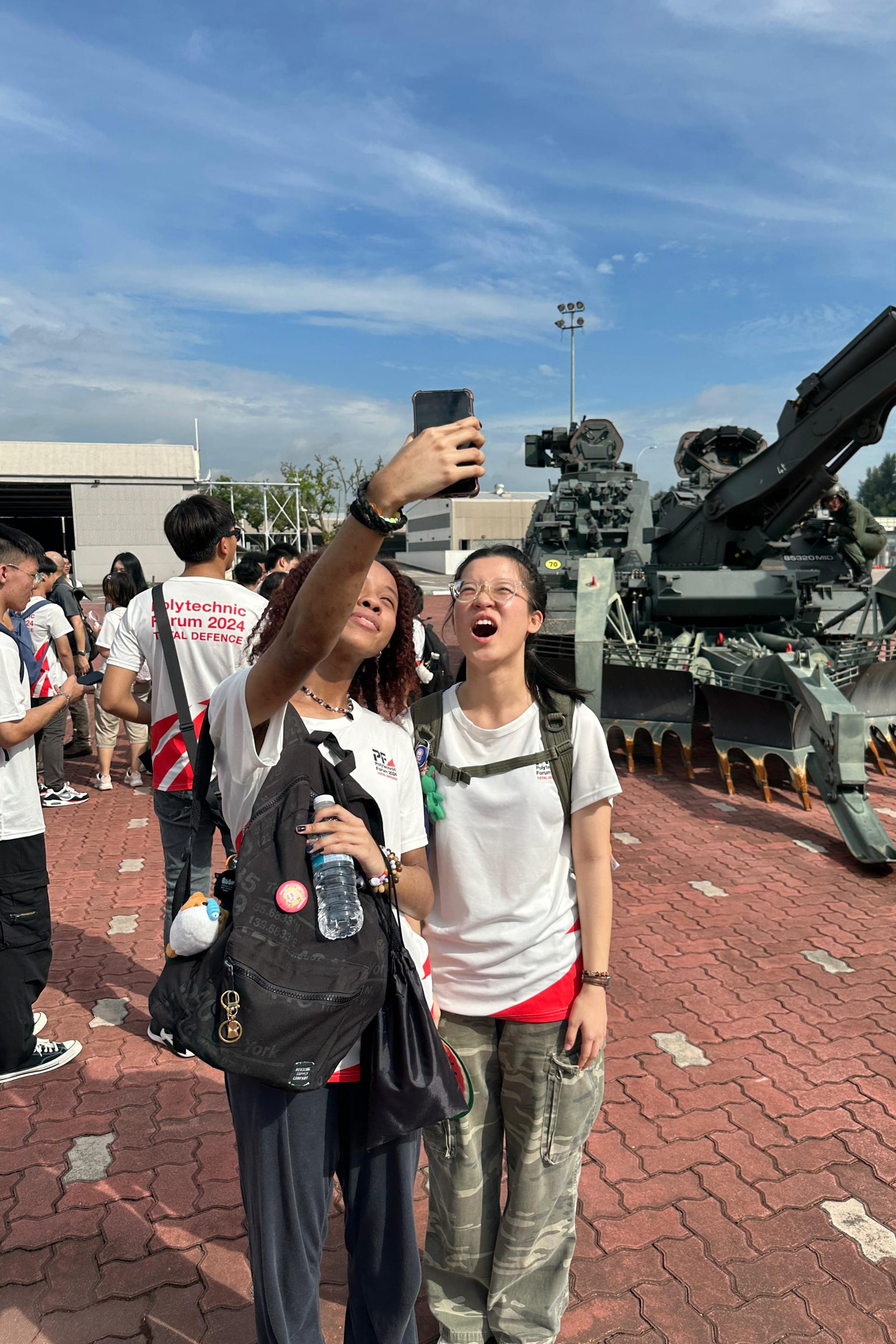 Two young women take a selfie outdoors near a large military vehicle at a Polytechnic Forum 2024 event.