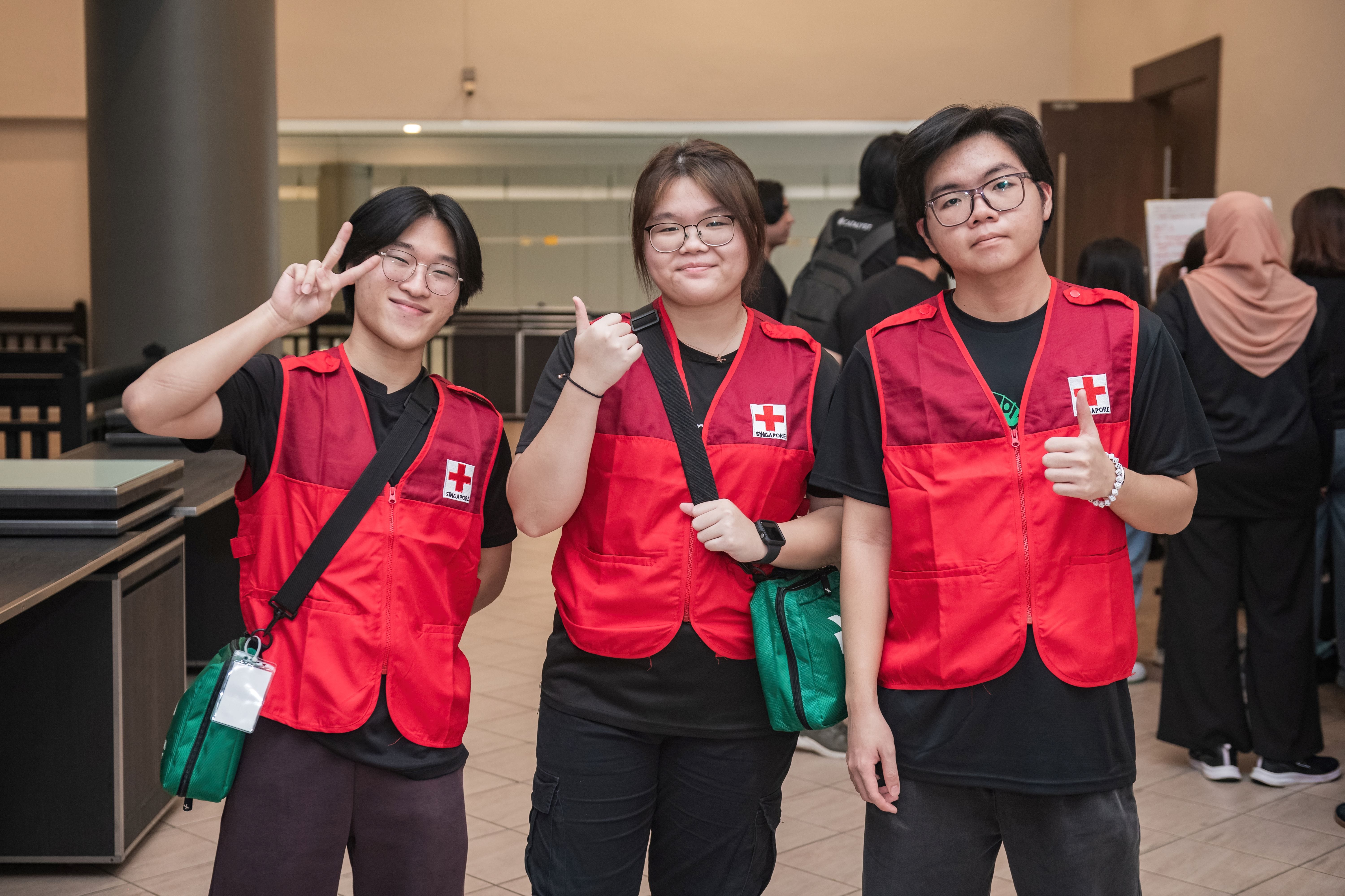 Three young Singapore Red Cross volunteers in red vests smile and pose indoors, one flashing a peace sign and one a thumbs up.