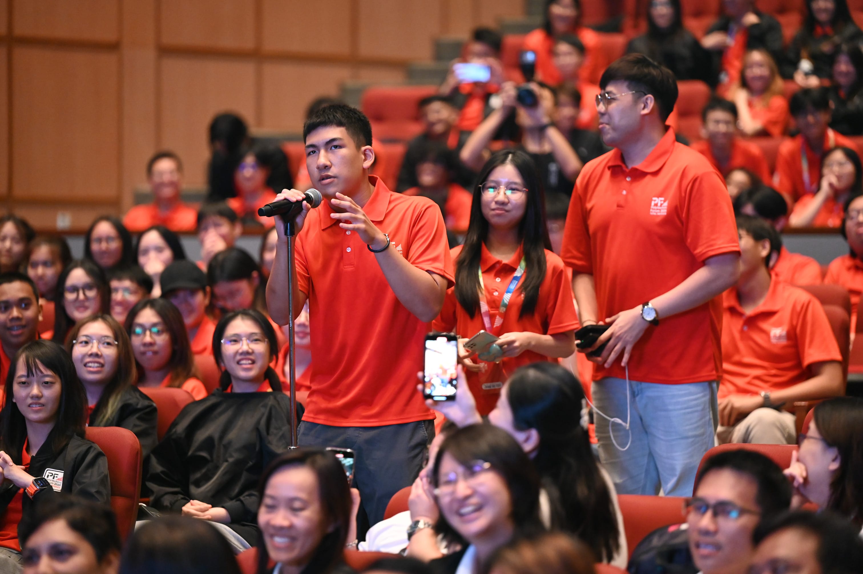 A young man in a red polo shirt speaks into a microphone in a packed auditorium filled with people in red shirts.
