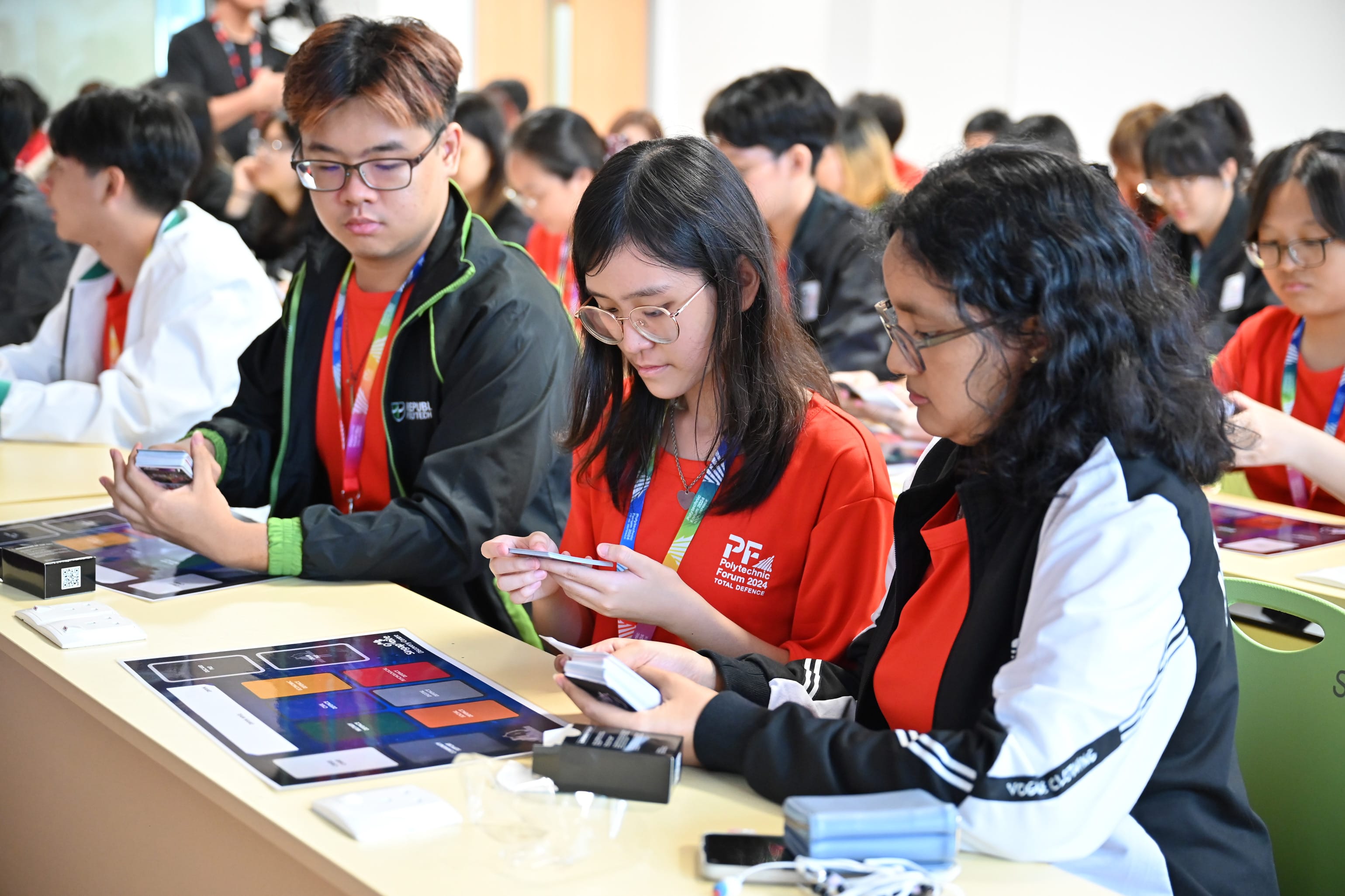 Students in red Polytechnic Forum shirts sit at tables, looking at cards and small devices during a group activity.