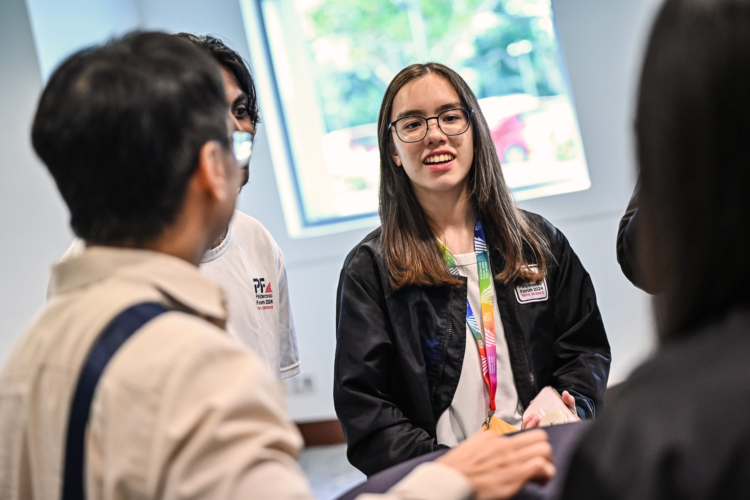 A smiling young woman with glasses and a colorful lanyard talks with a small group at a Polytechnic Forum 2024 event.
