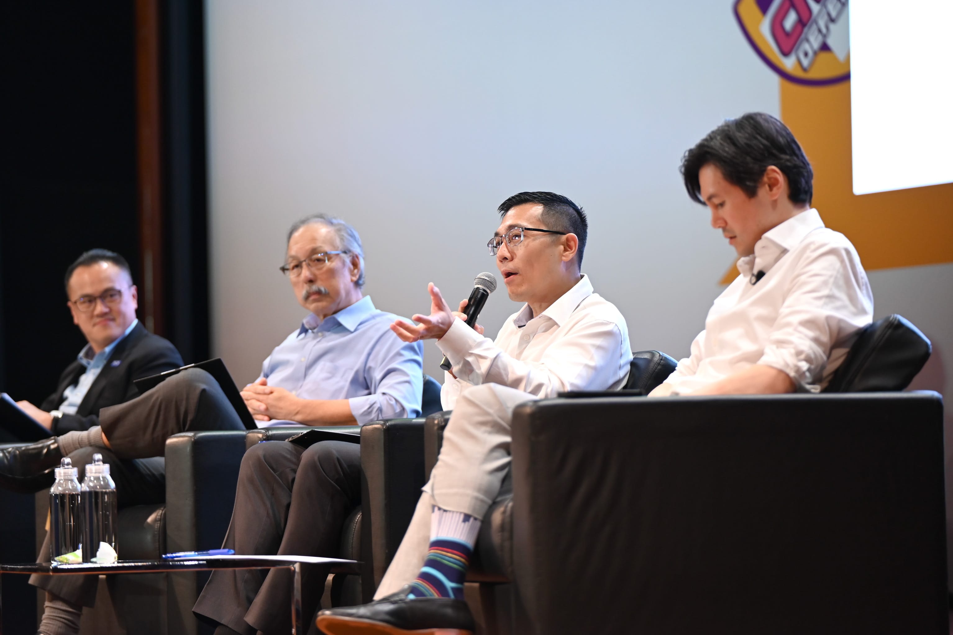 Four men sit in black chairs on a stage; one holds a microphone and gestures while speaking at a panel event.