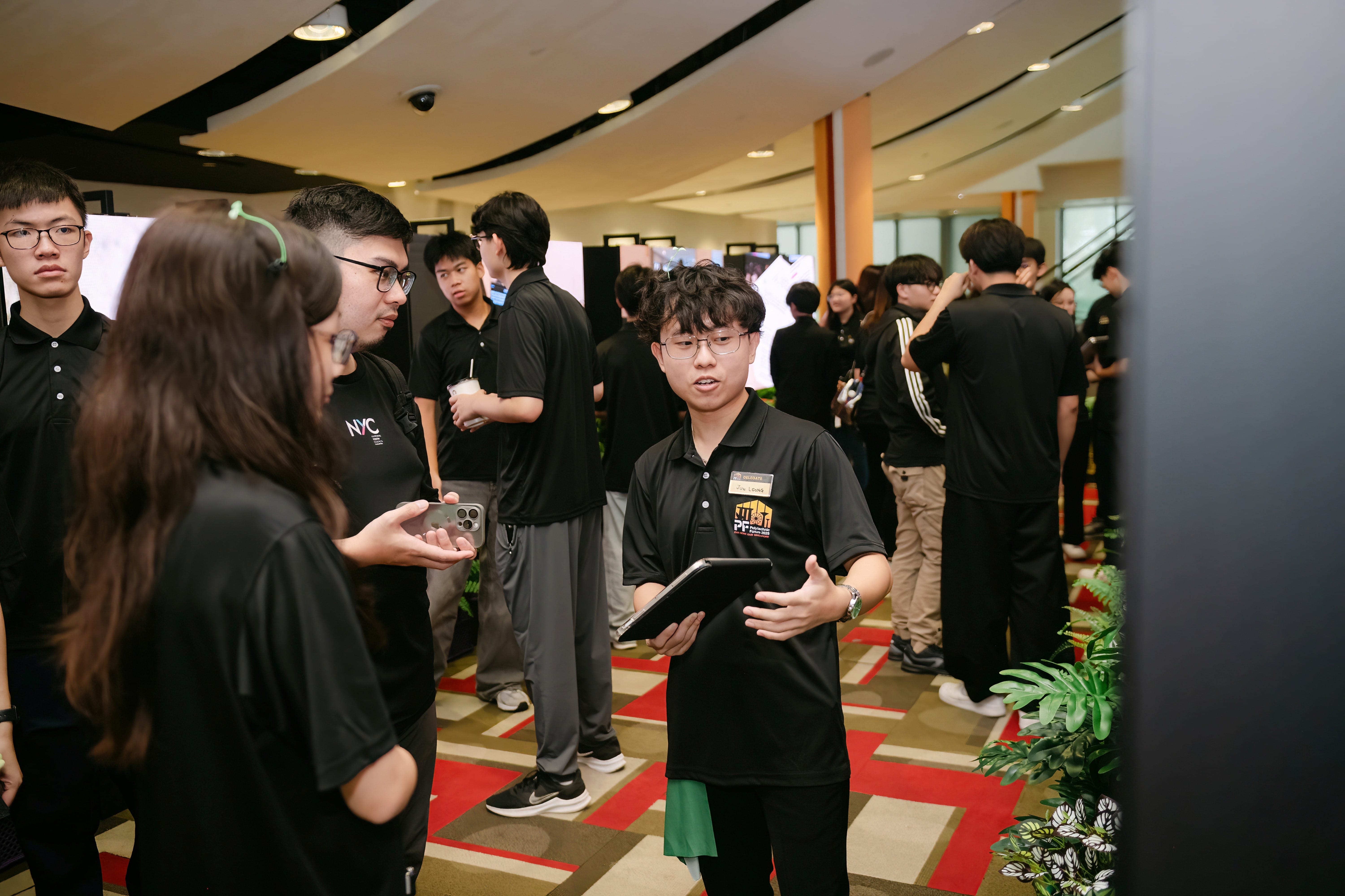 Young people in black polo shirts gather indoors; one holds a tablet and speaks to two others at an event.