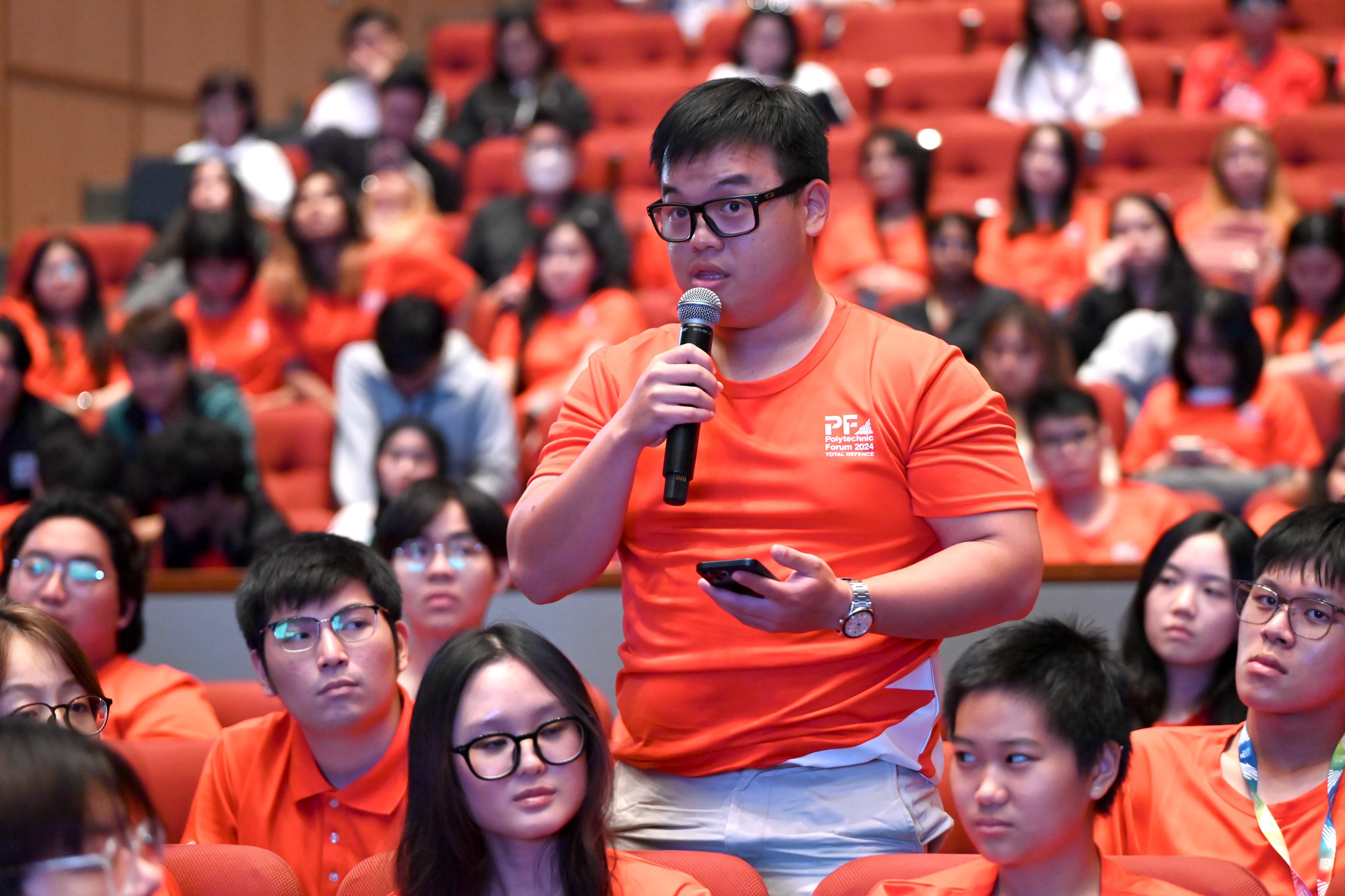 A young man in an orange shirt speaks into a microphone in a packed auditorium of students wearing orange shirts.
