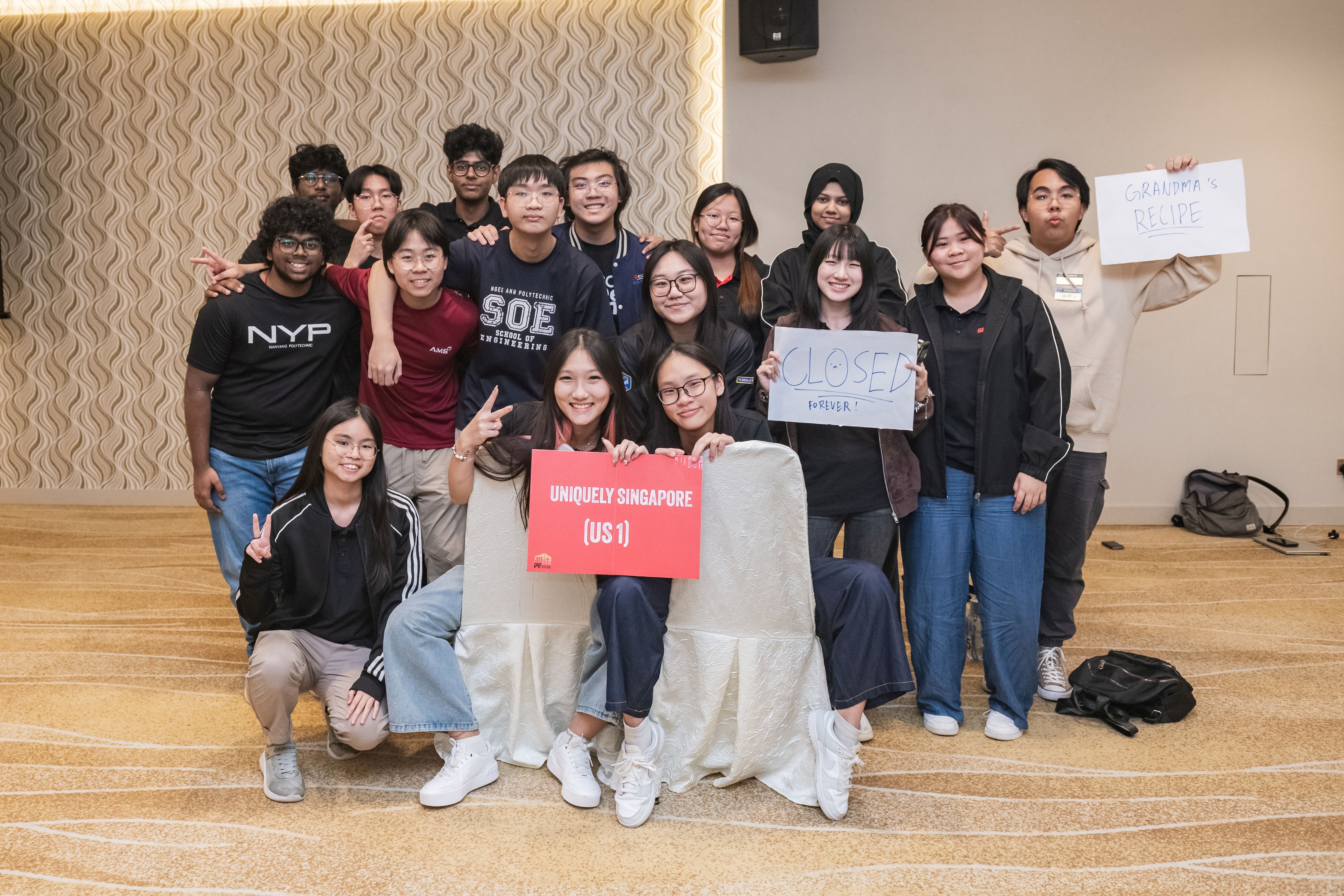 A group of students posing together, holding signs reading "Uniquely Singapore US1," "Closed Forever," and "Grandma's Recipe."