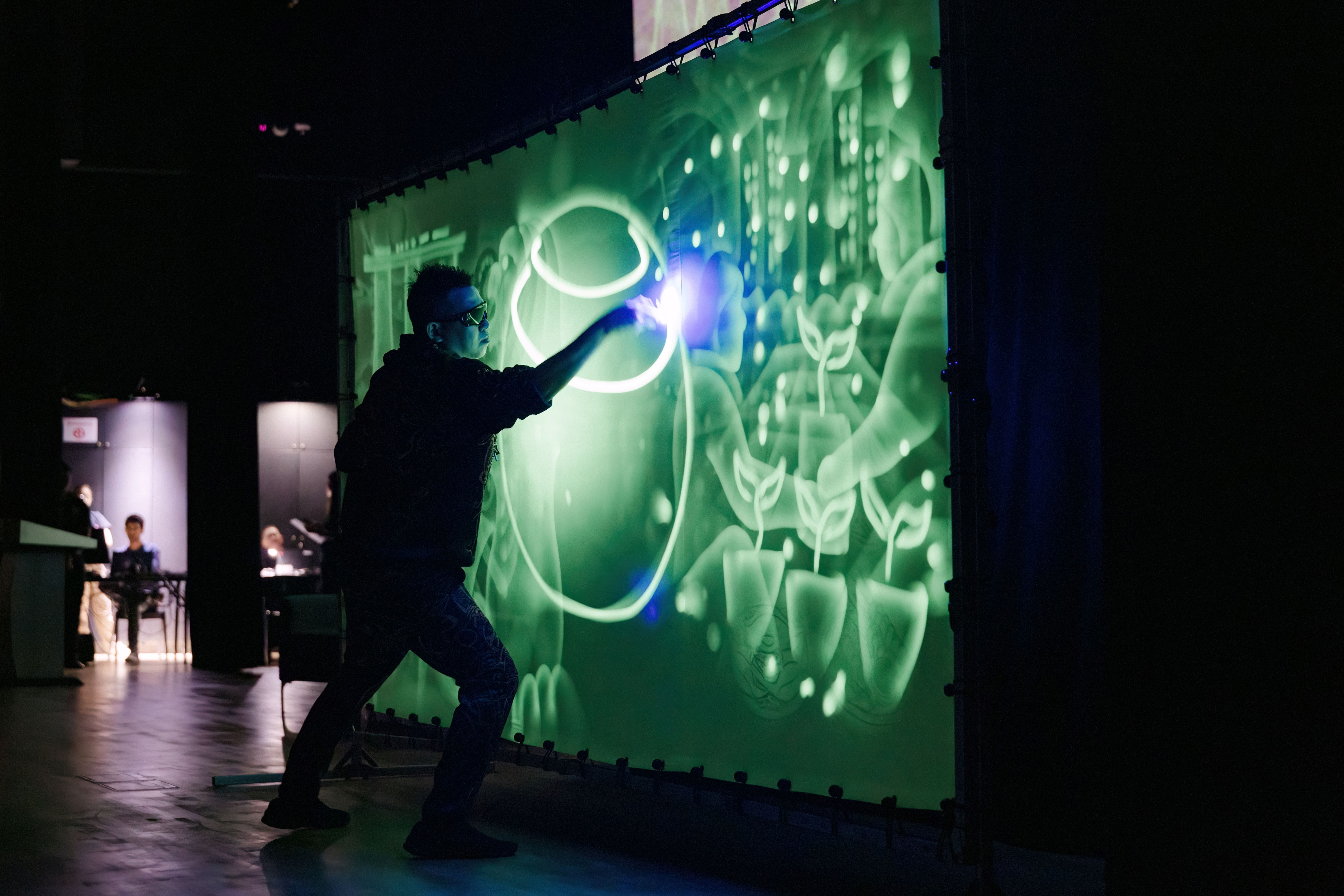 A person in dark clothing touches a large green illuminated projection screen showing glowing rings and plant designs.