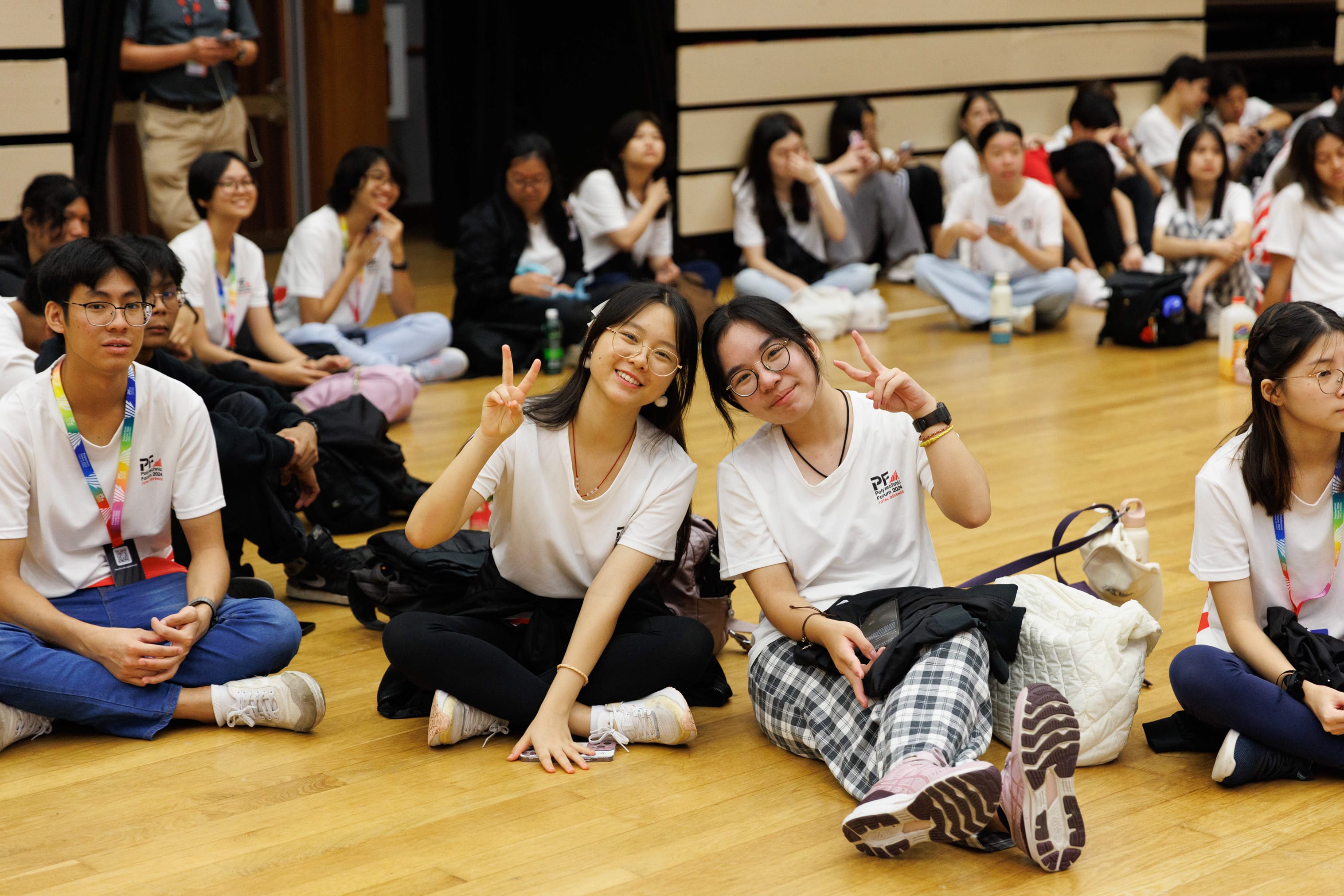 Two smiling young women in white shirts flash peace signs while sitting on a wooden floor among a large group of students.