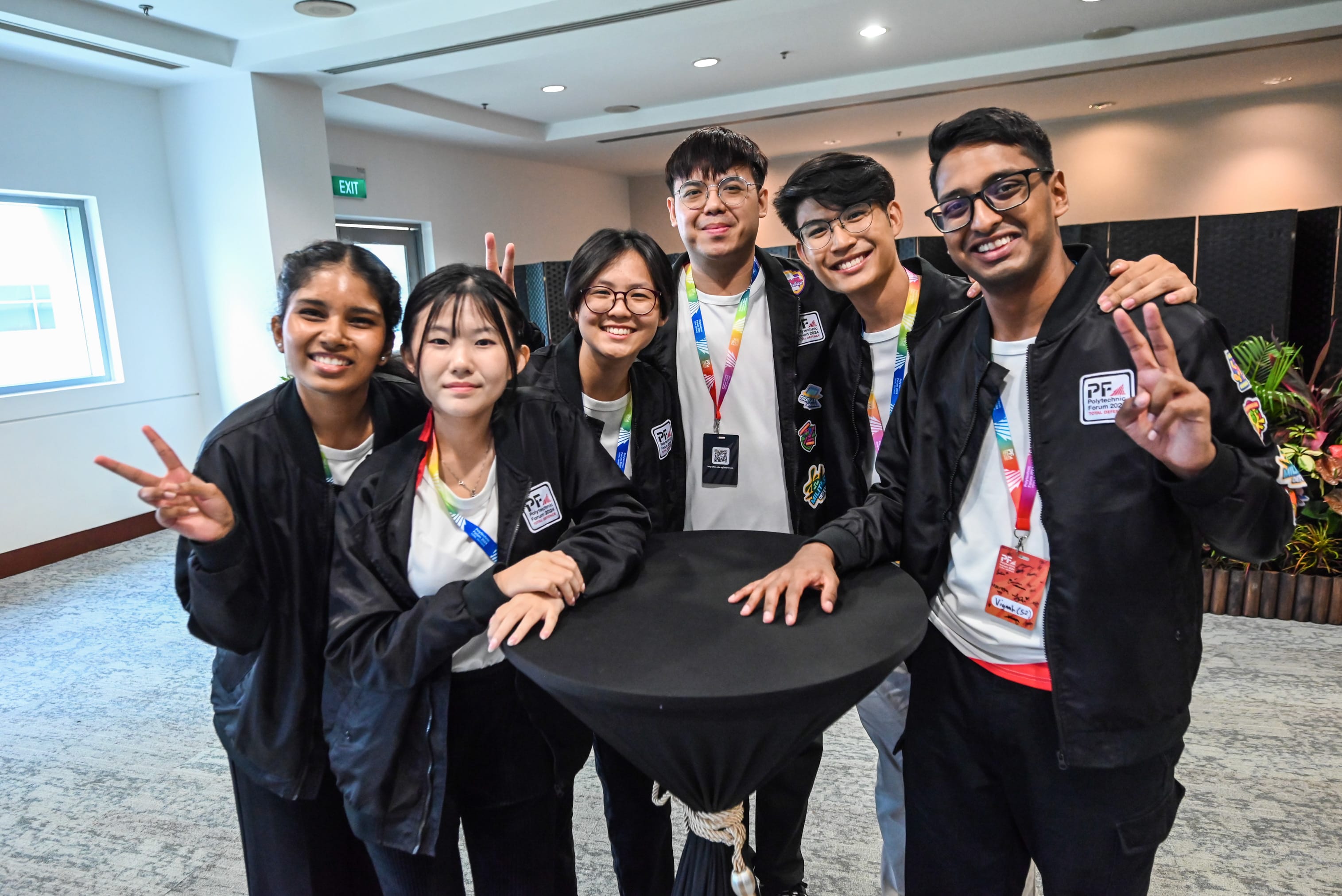Six smiling young people in black Polytechnic Forum jackets pose together around a tall round table indoors.