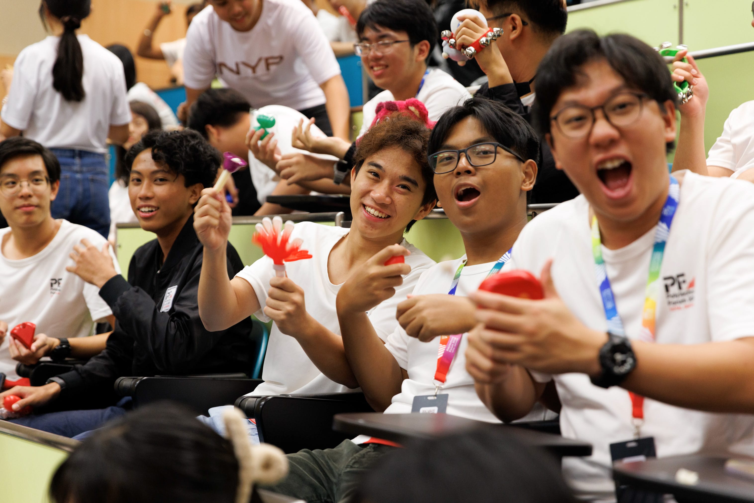 Young students in white shirts laugh and hold colorful small instruments like maracas during a lively group activity.